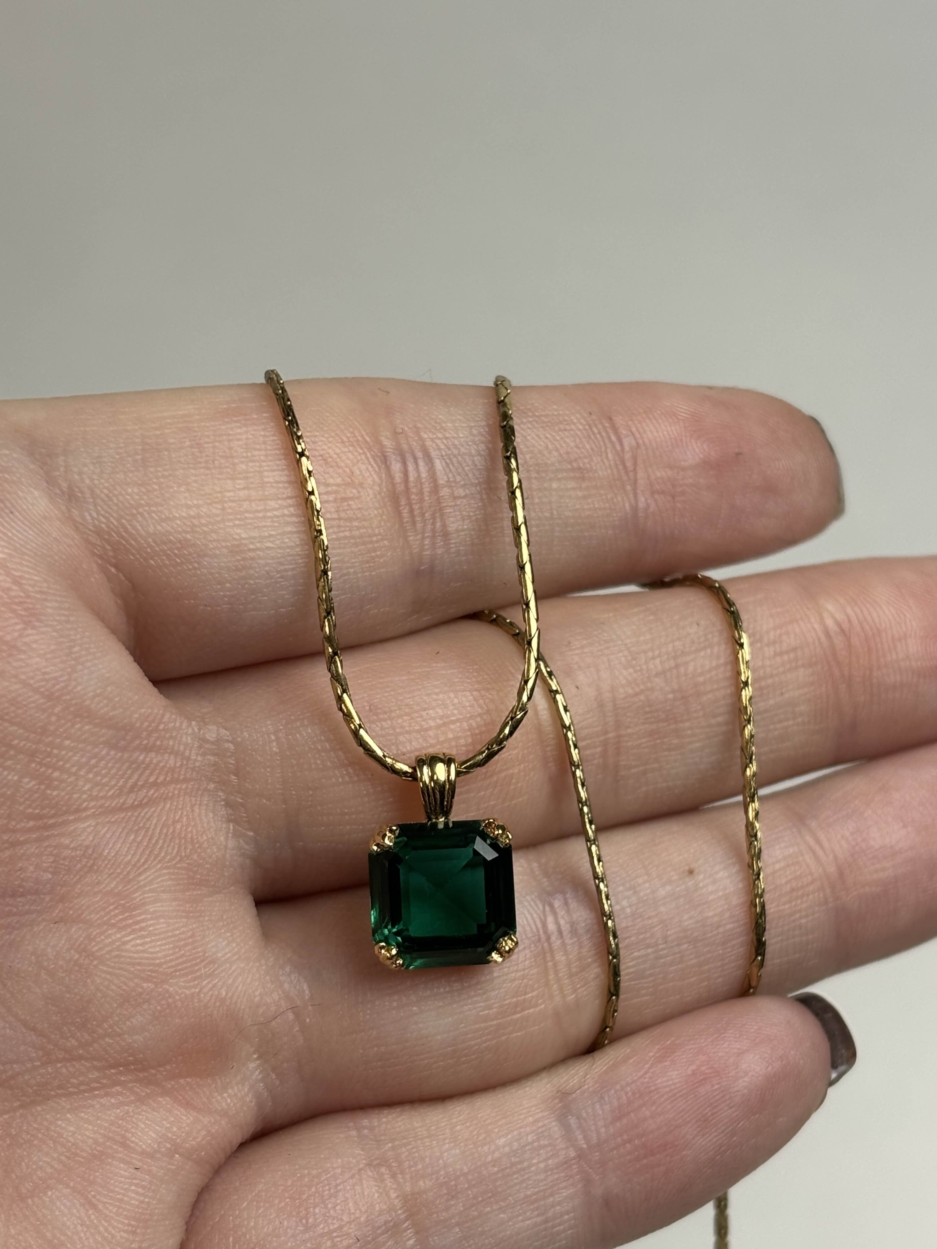 Front of a gold necklace with a green gemstone held in a hand against a neutral background