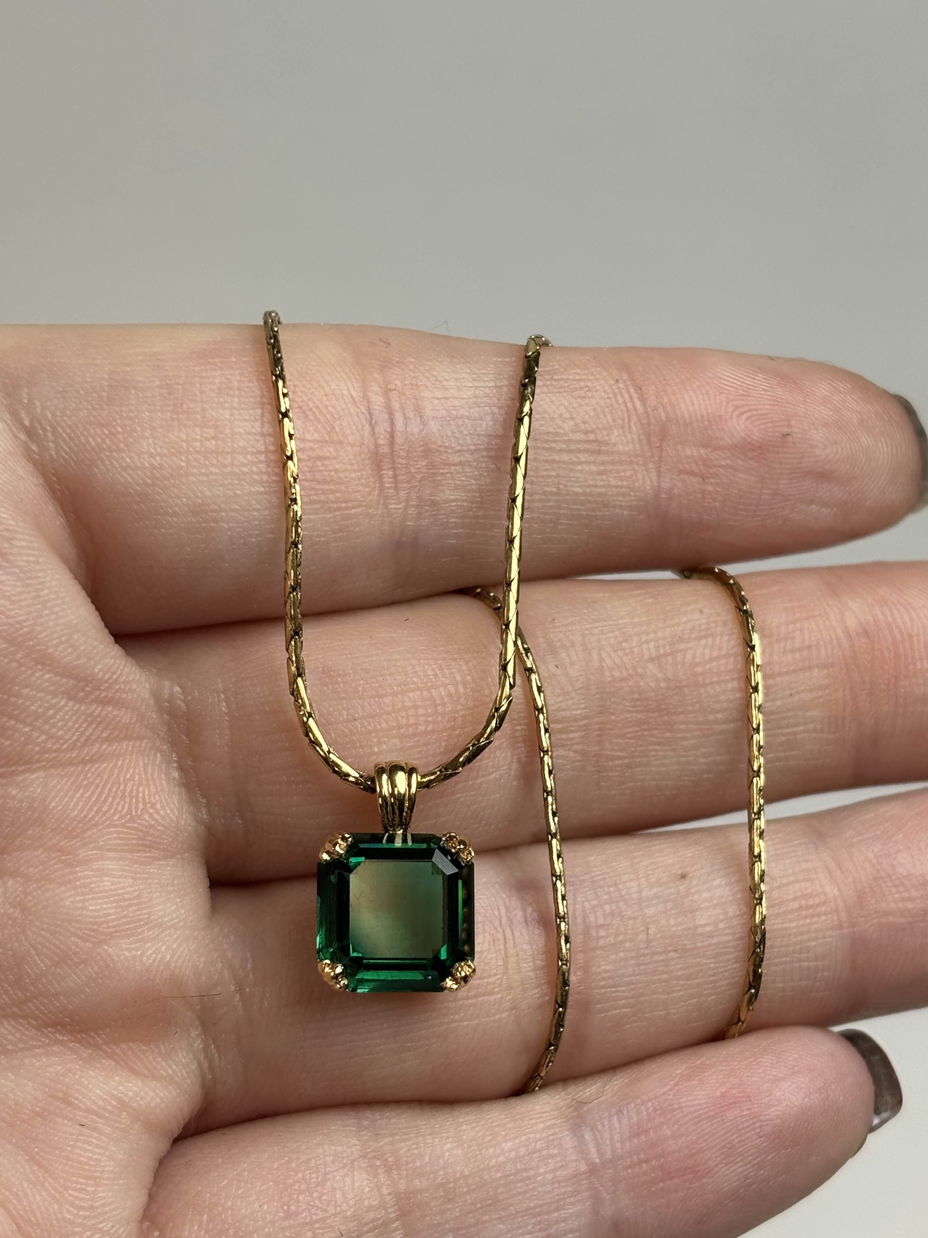 Front of a necklace with a green gemstone pendant held on a hand against a neutral background