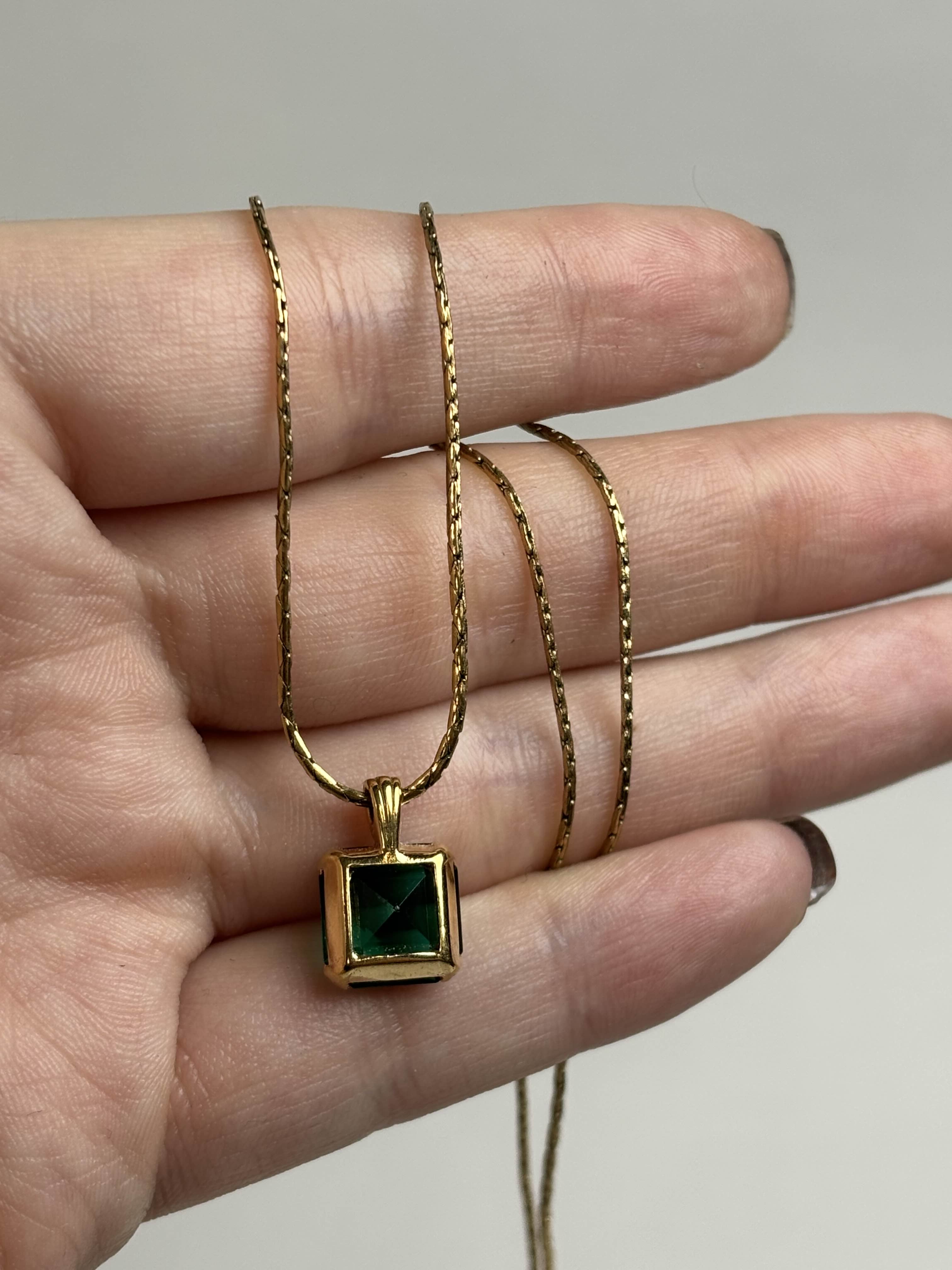 Back of a gold necklace with a green gemstone held in a hand against a neutral background