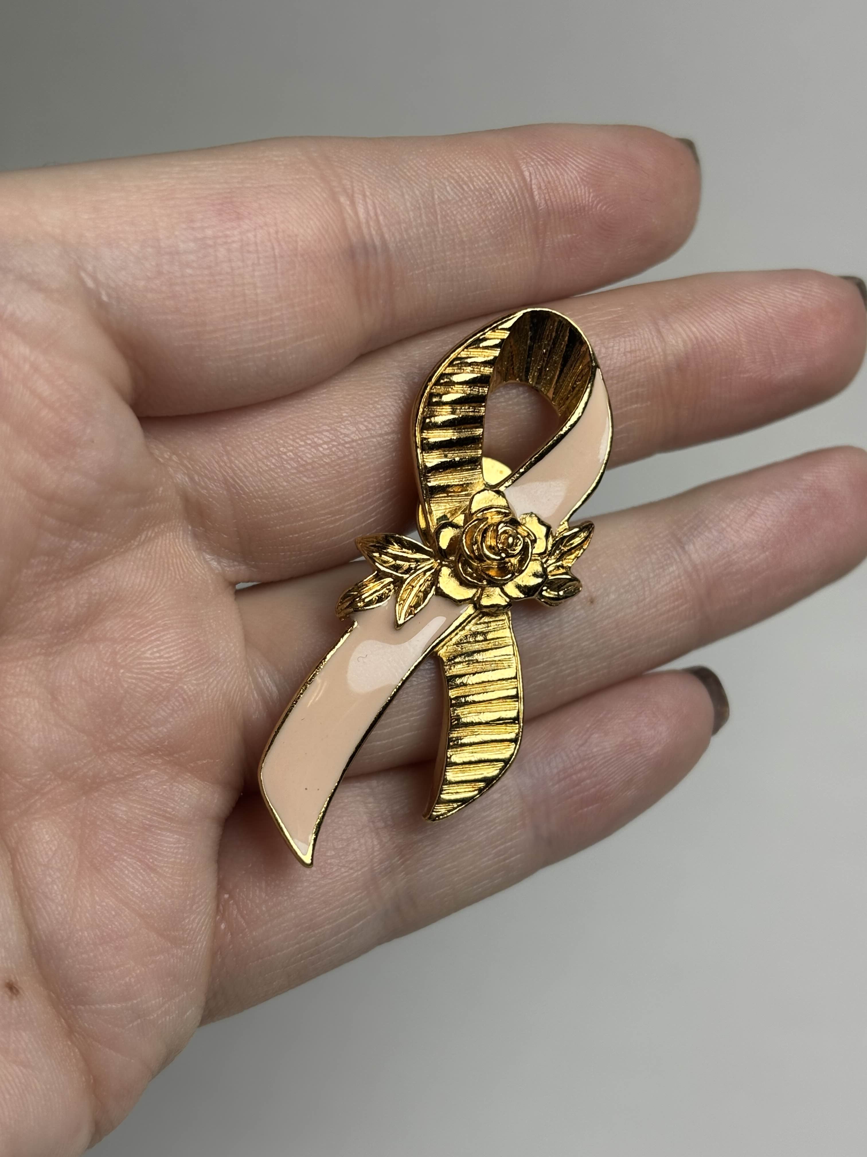 Front of breast cancer awareness ribbon brooch with pink enamel and a gold rose in the center held in a hand against a neutral background