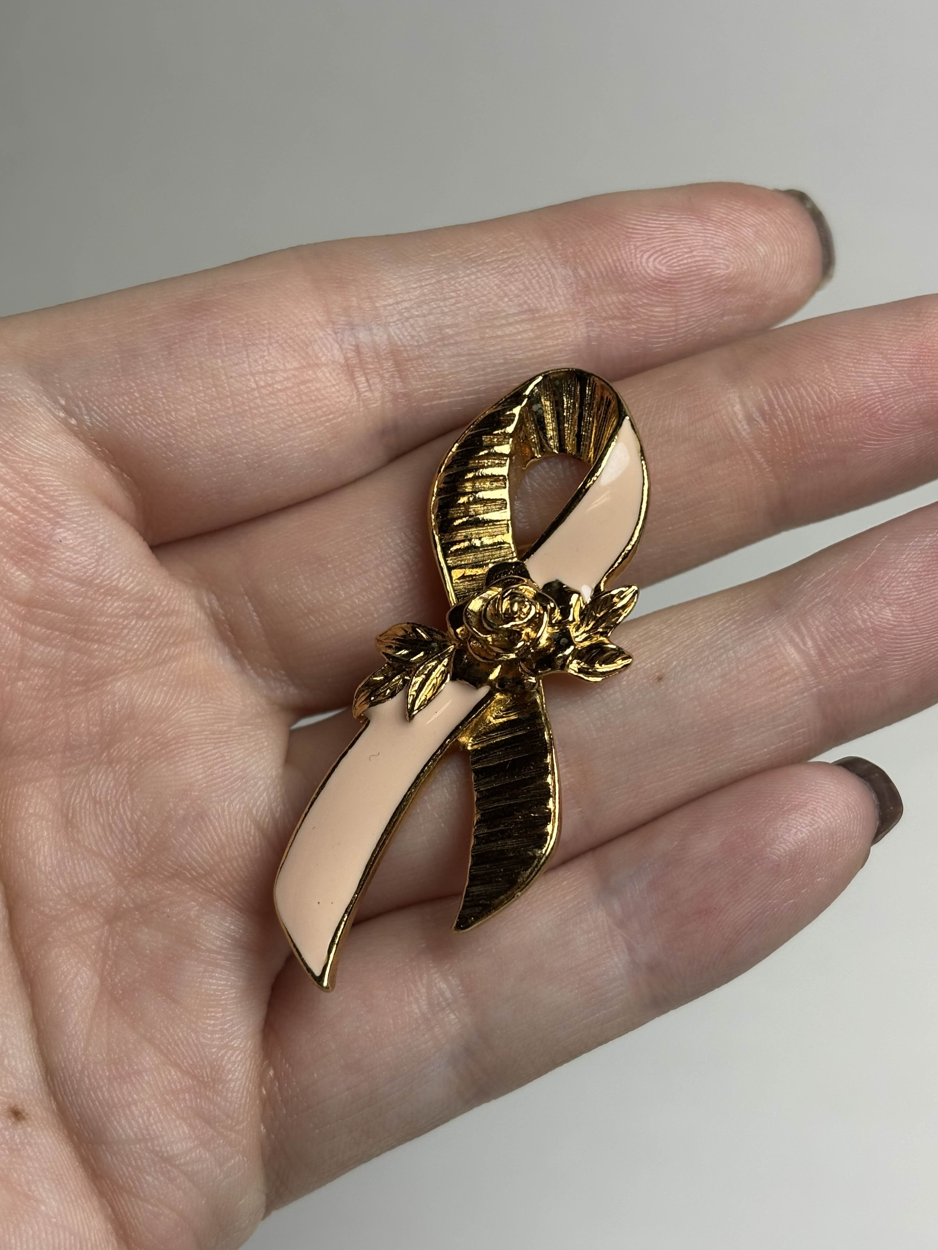 Front of breast cancer awareness ribbon brooch with pink enamel and a gold rose in the center held in a hand against a neutral background