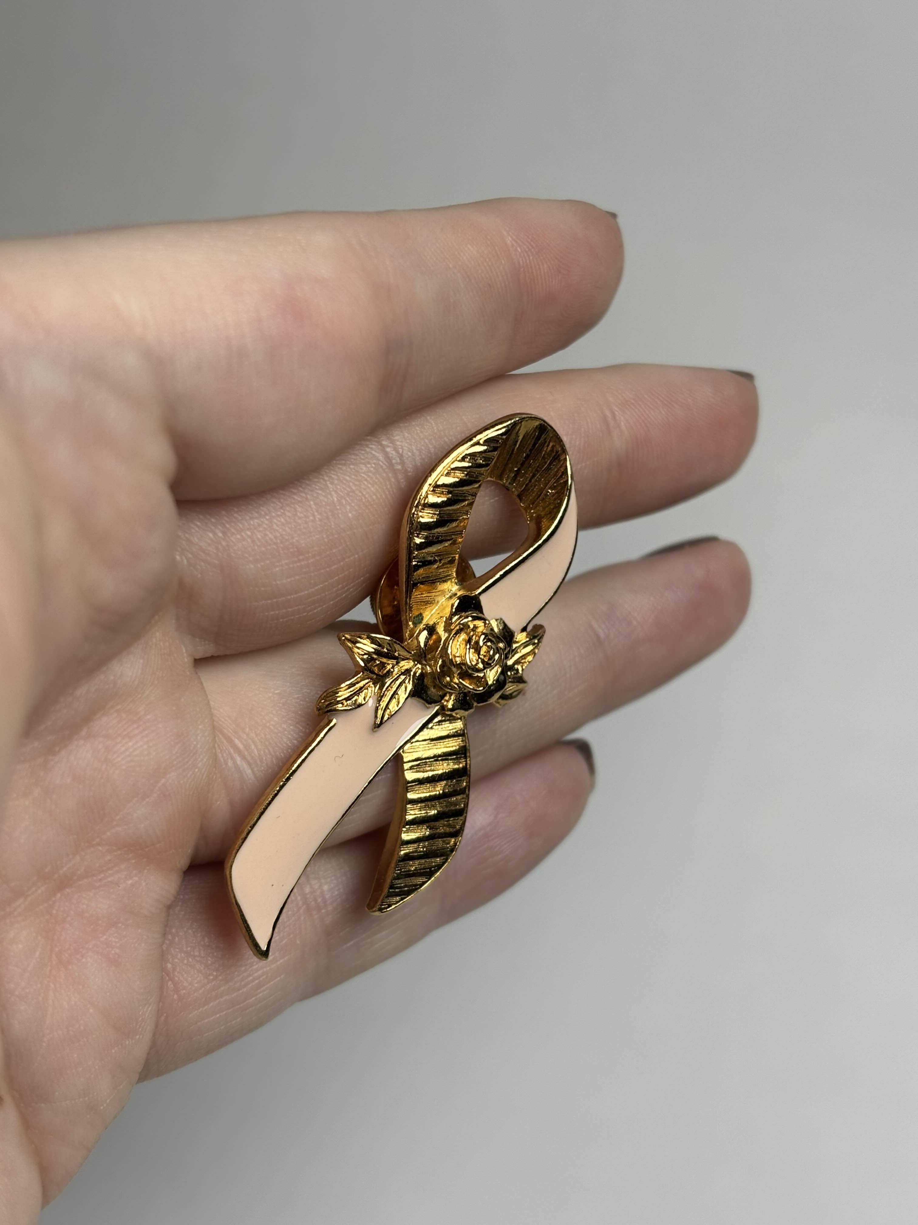 Front of breast cancer awareness ribbon brooch with pink enamel and a gold rose in the center held in a hand against a neutral background