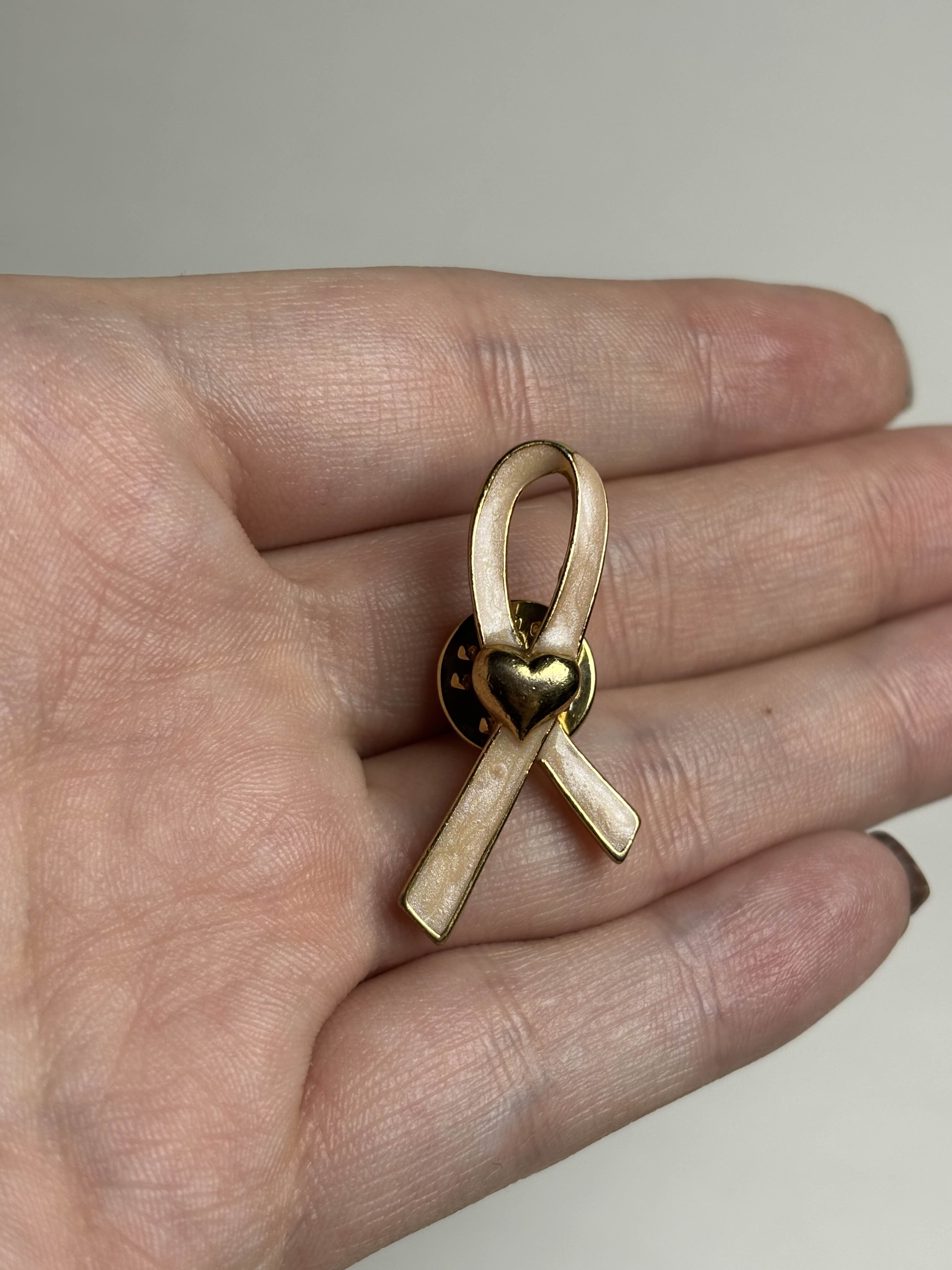 Front of gold breast cancer awareness ribbon-shaped brooch with pink enamel and a heart in the middle on a hand against a neutral background