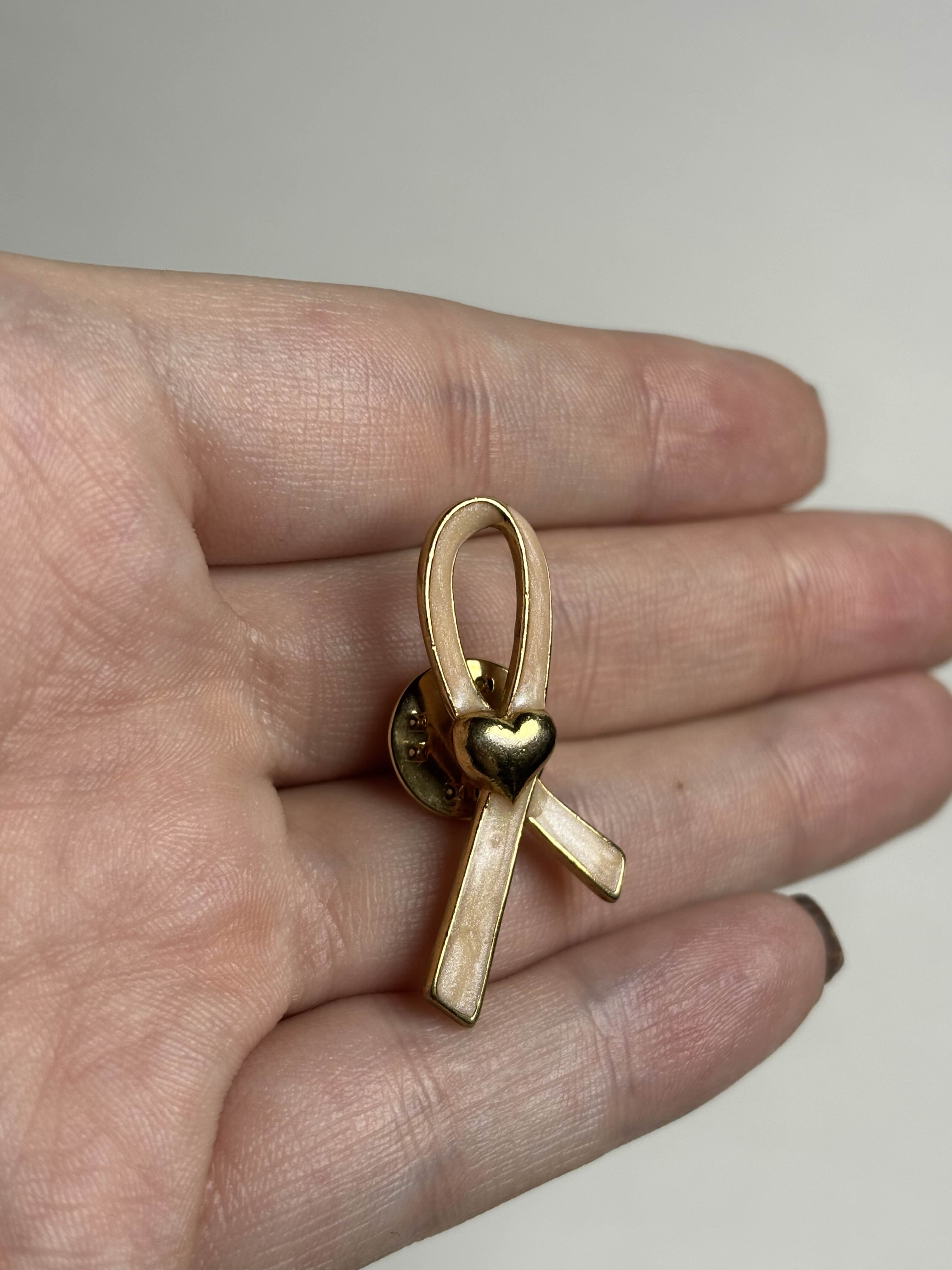 Front of gold breast cancer awareness ribbon-shaped brooch with pink enamel and a heart in the middle on a hand against a neutral background