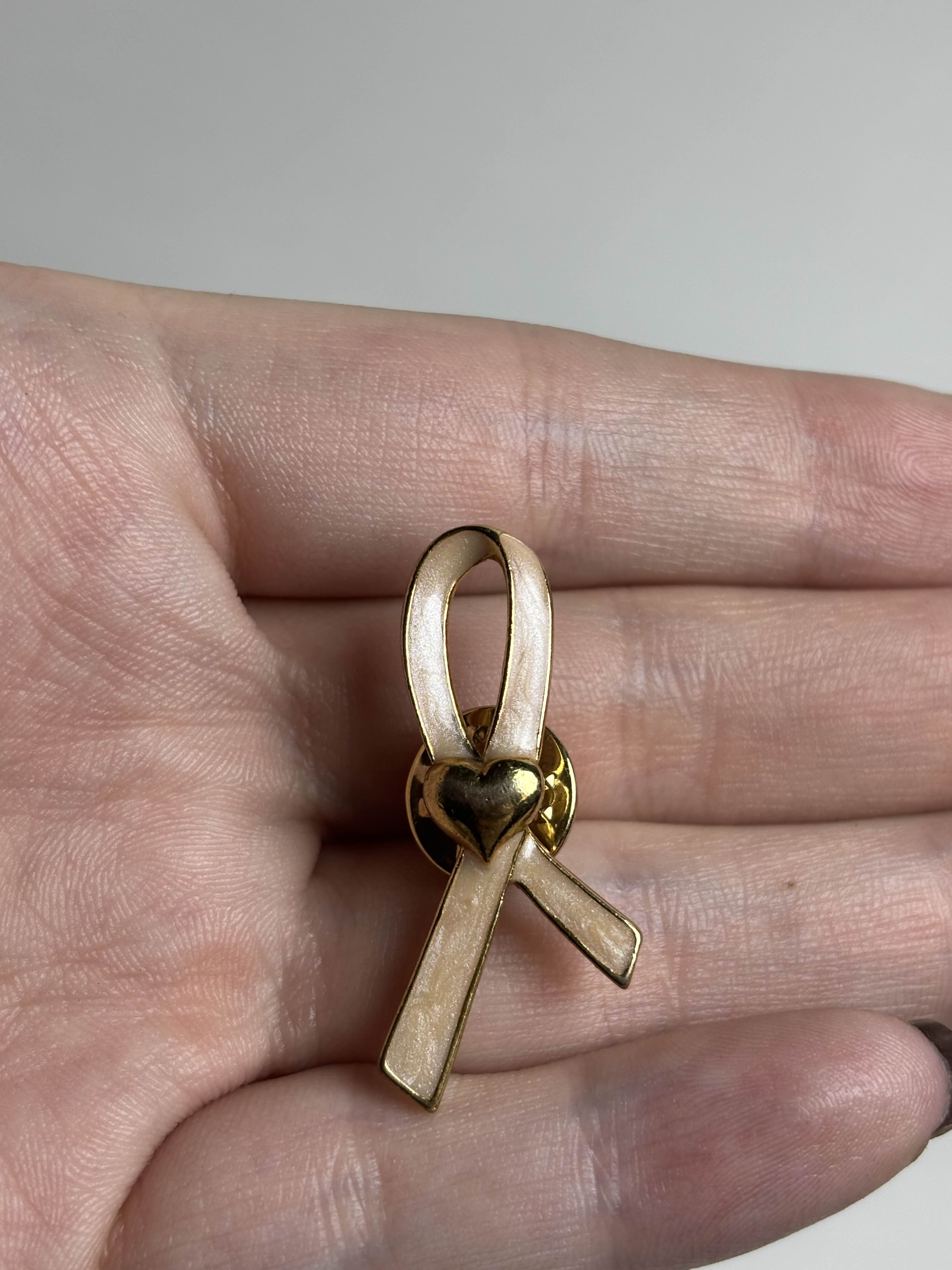 Front of gold breast cancer awareness ribbon-shaped brooch with pink enamel and a heart in the middle on a hand against a neutral background