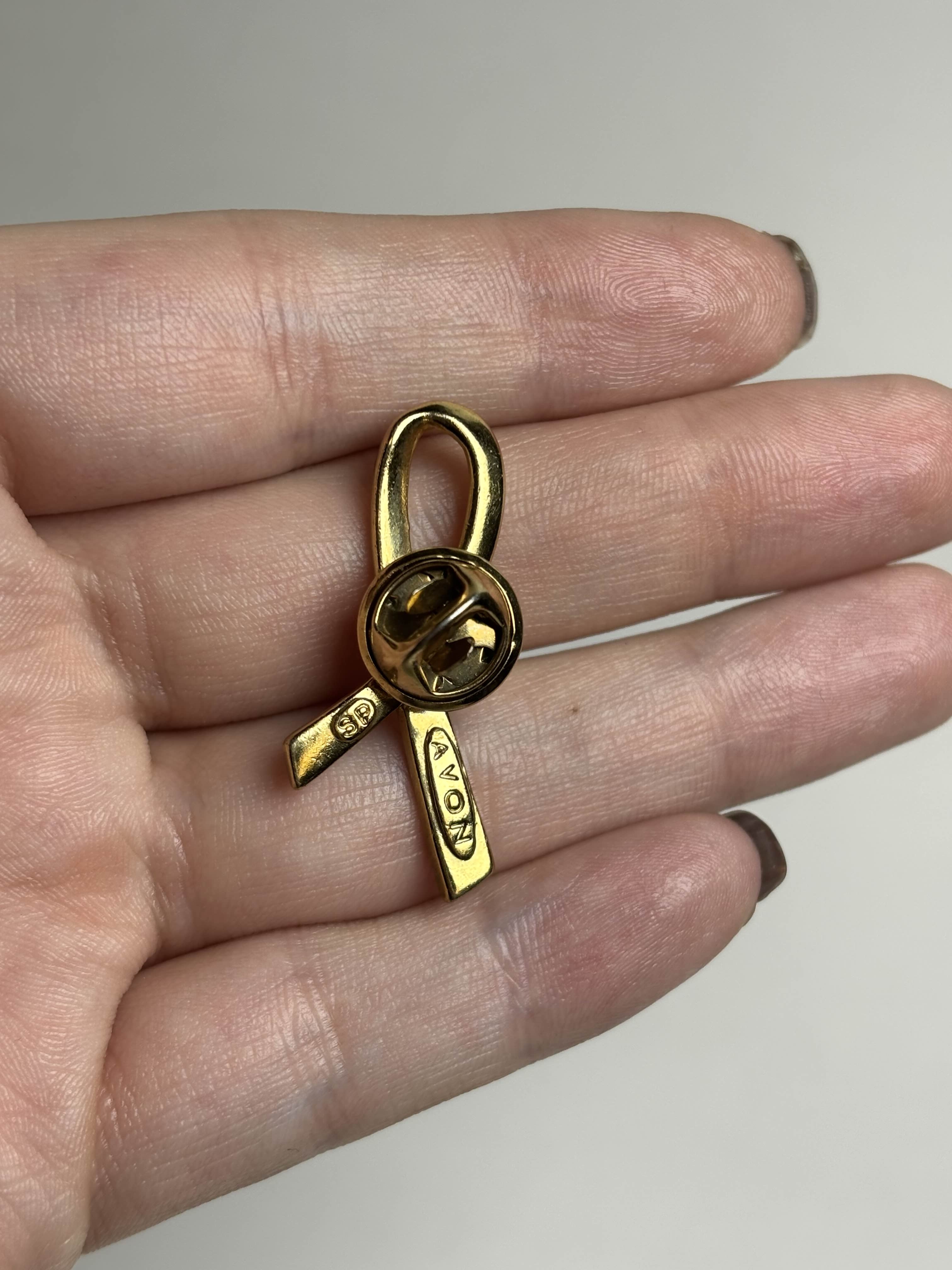 Back of gold breast cancer awareness ribbon-shaped brooch on a hand against a neutral background