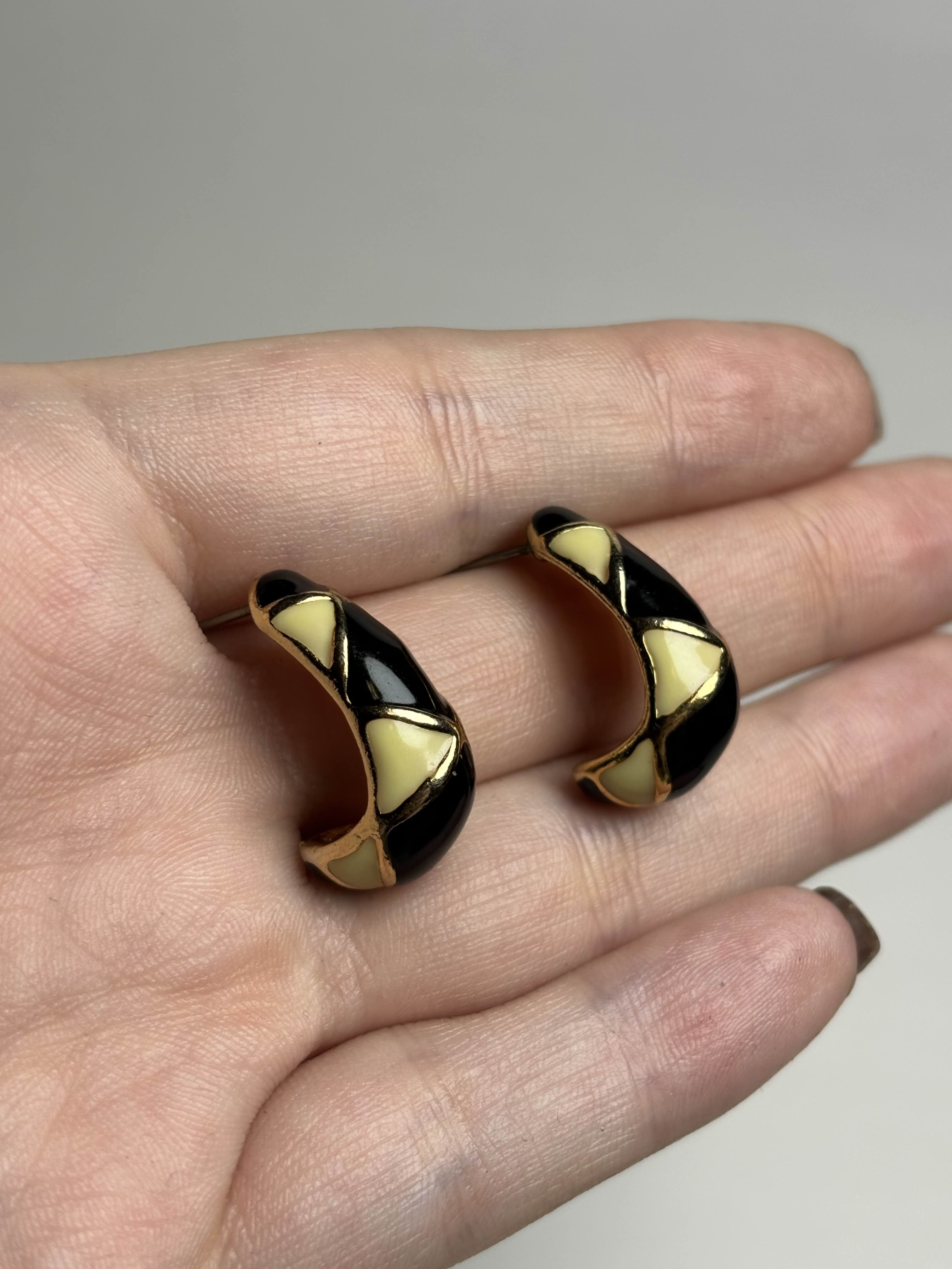 Front of pair of black, cream and gold earrings held between fingers on a neutral background