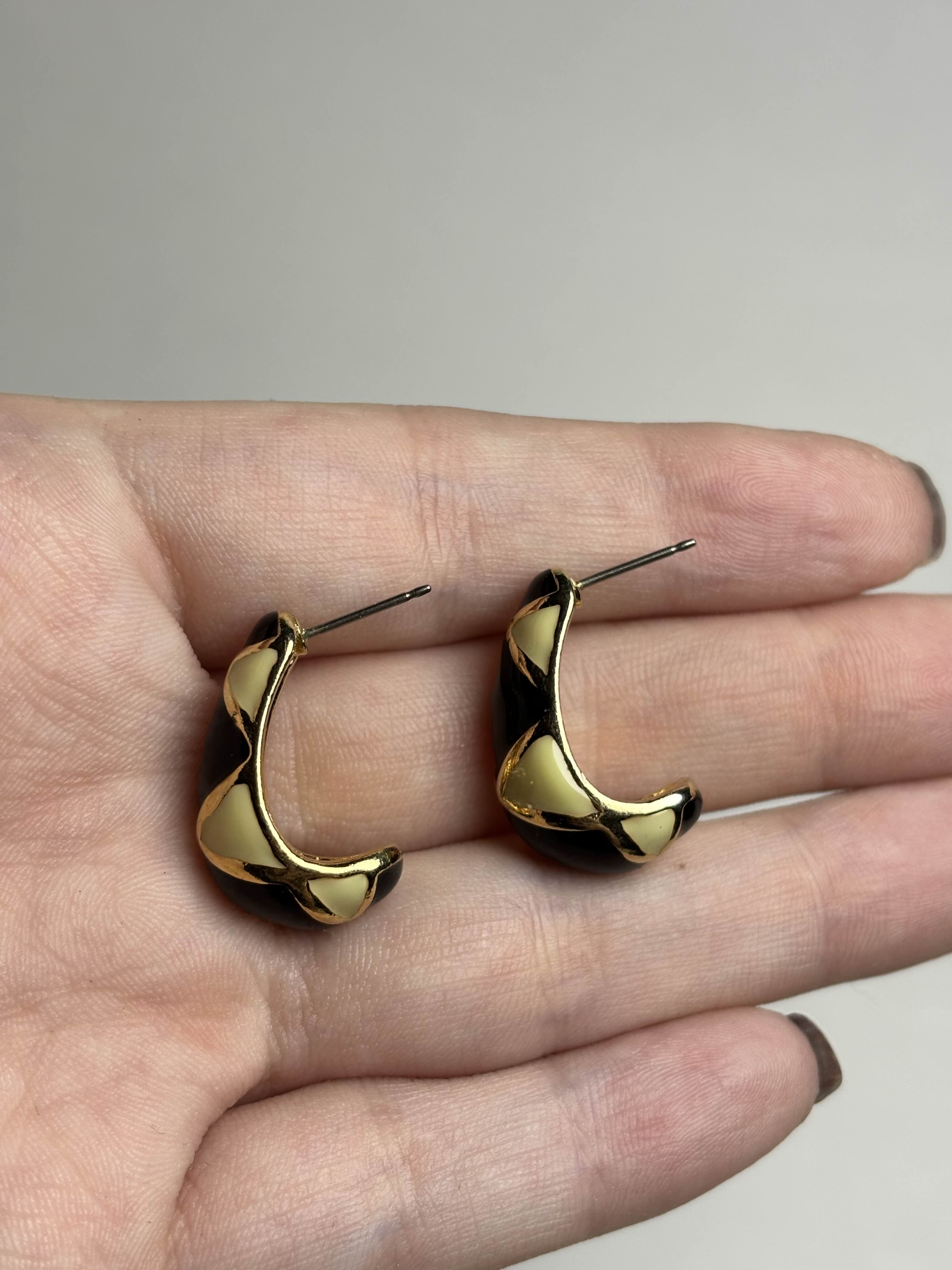 Front of pair of black, cream and gold earrings held between fingers on a neutral background