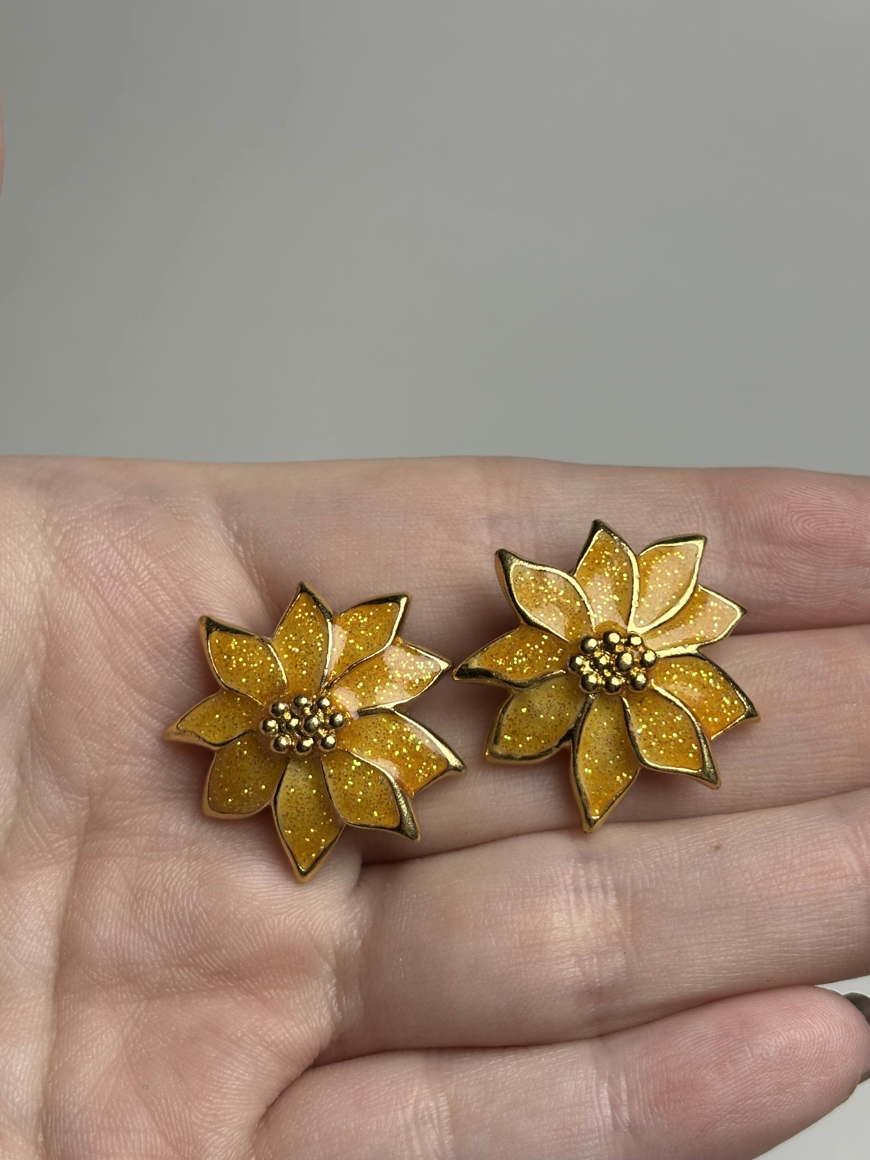 Front of gold and orange Poinsettia shaped earrings held in a hand against a neutral background