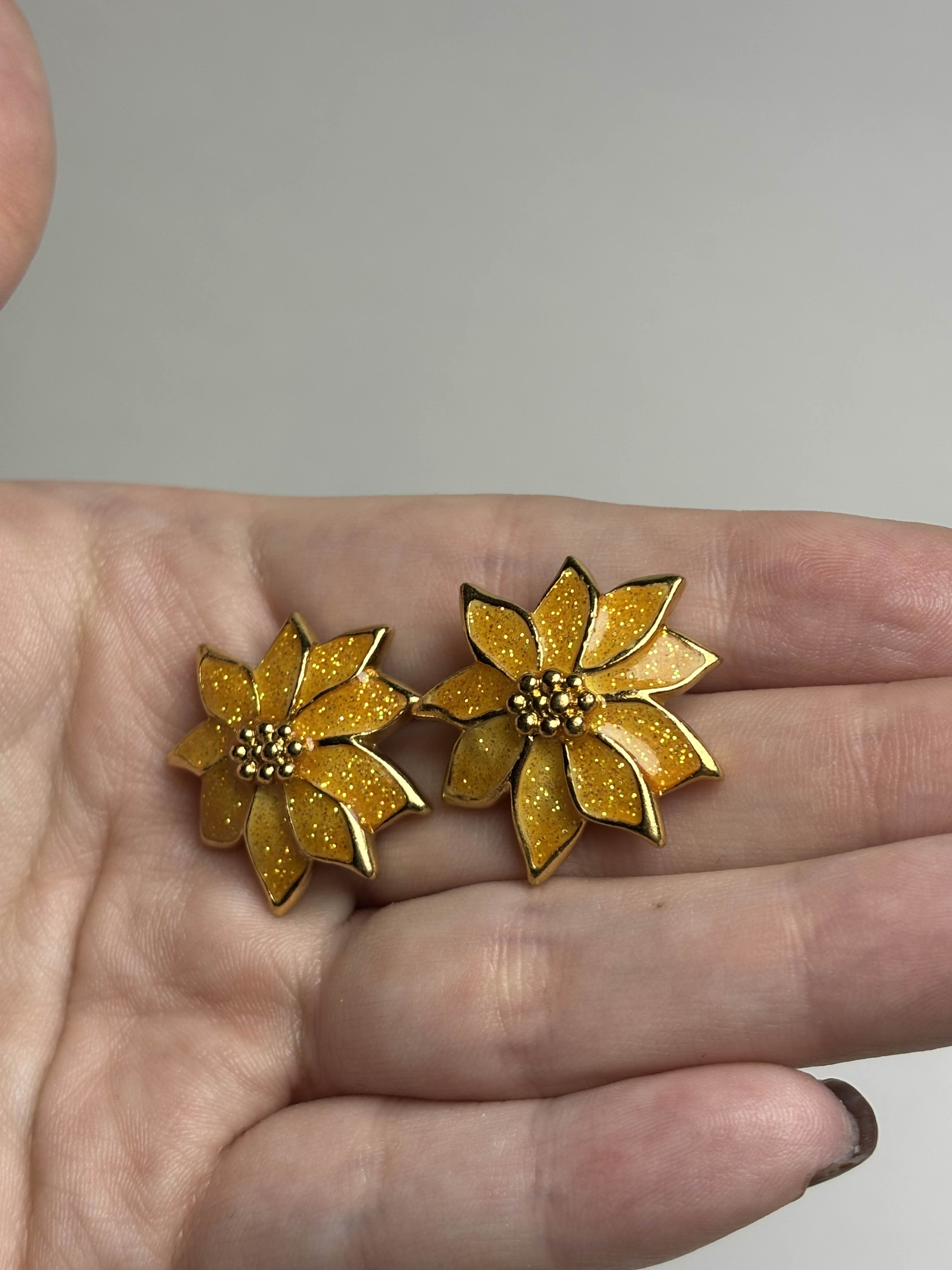 Front of gold and orange Poinsettia shaped earrings held in a hand against a neutral background