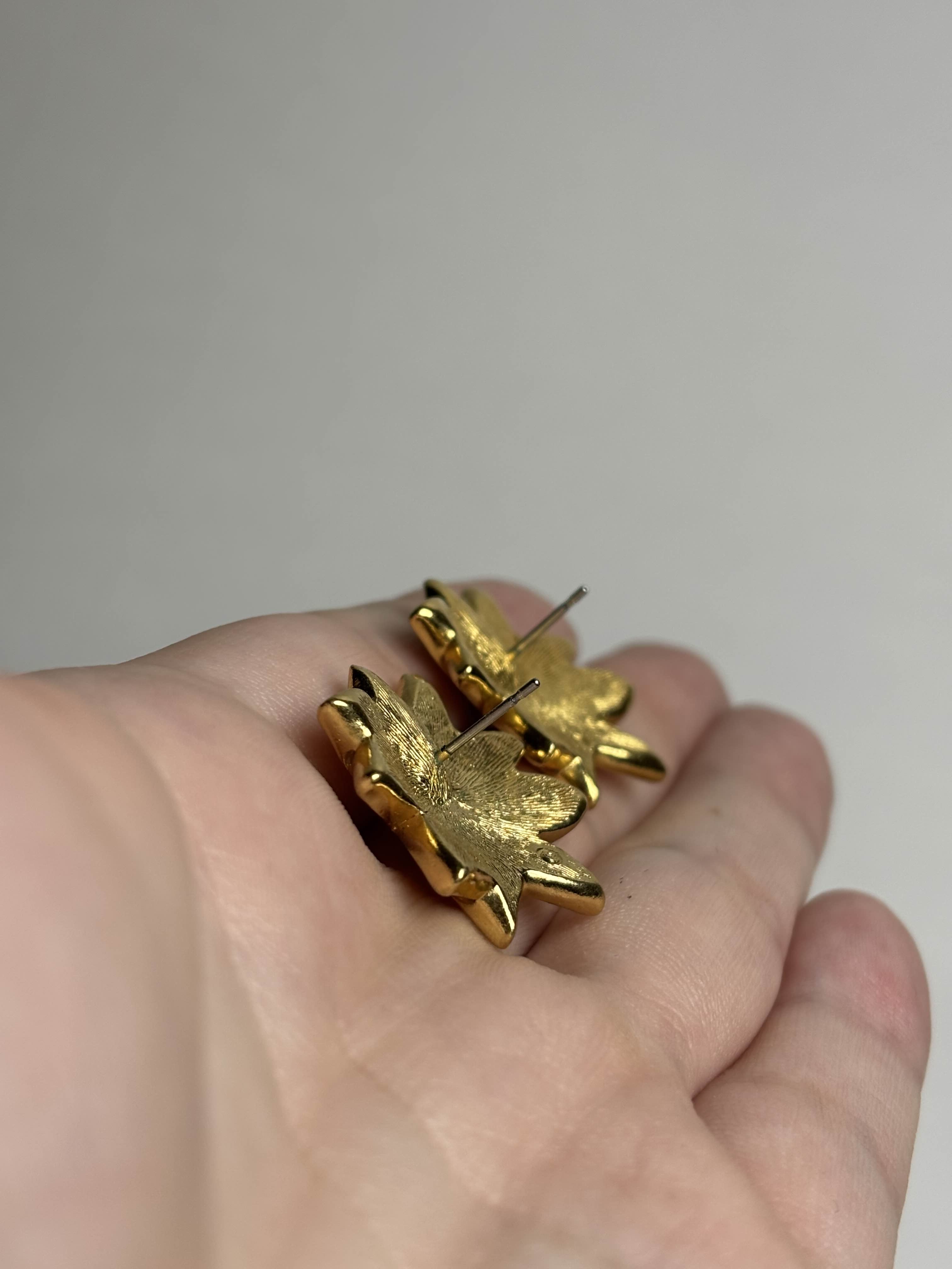 Front of gold and orange Poinsettia shaped earrings showing posts held in a hand against a neutral background
