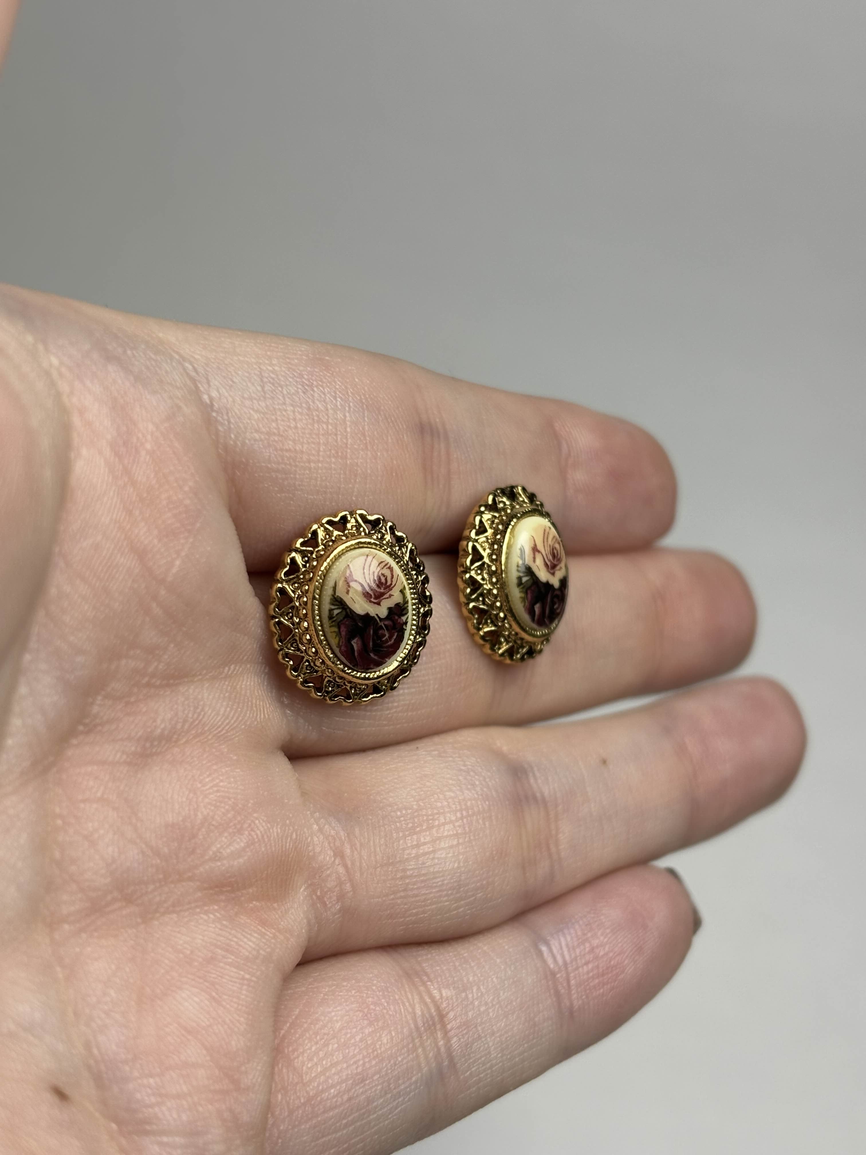 Front of oval earrings with rose design in center and gold filagree heart boarder held in a hand against a neutral background