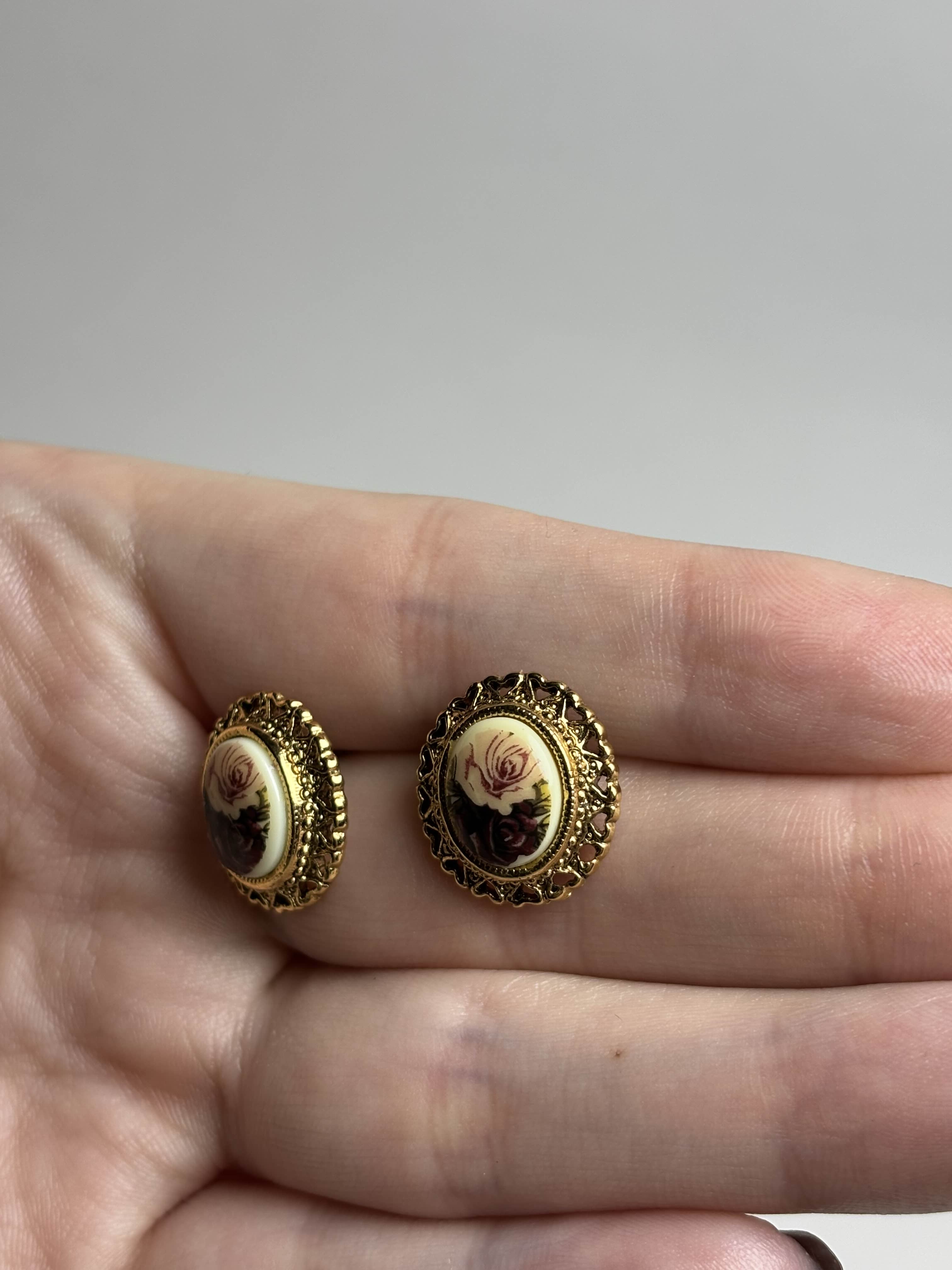 Front of oval earrings with rose design in center and gold filagree heart boarder held in a hand against a neutral background