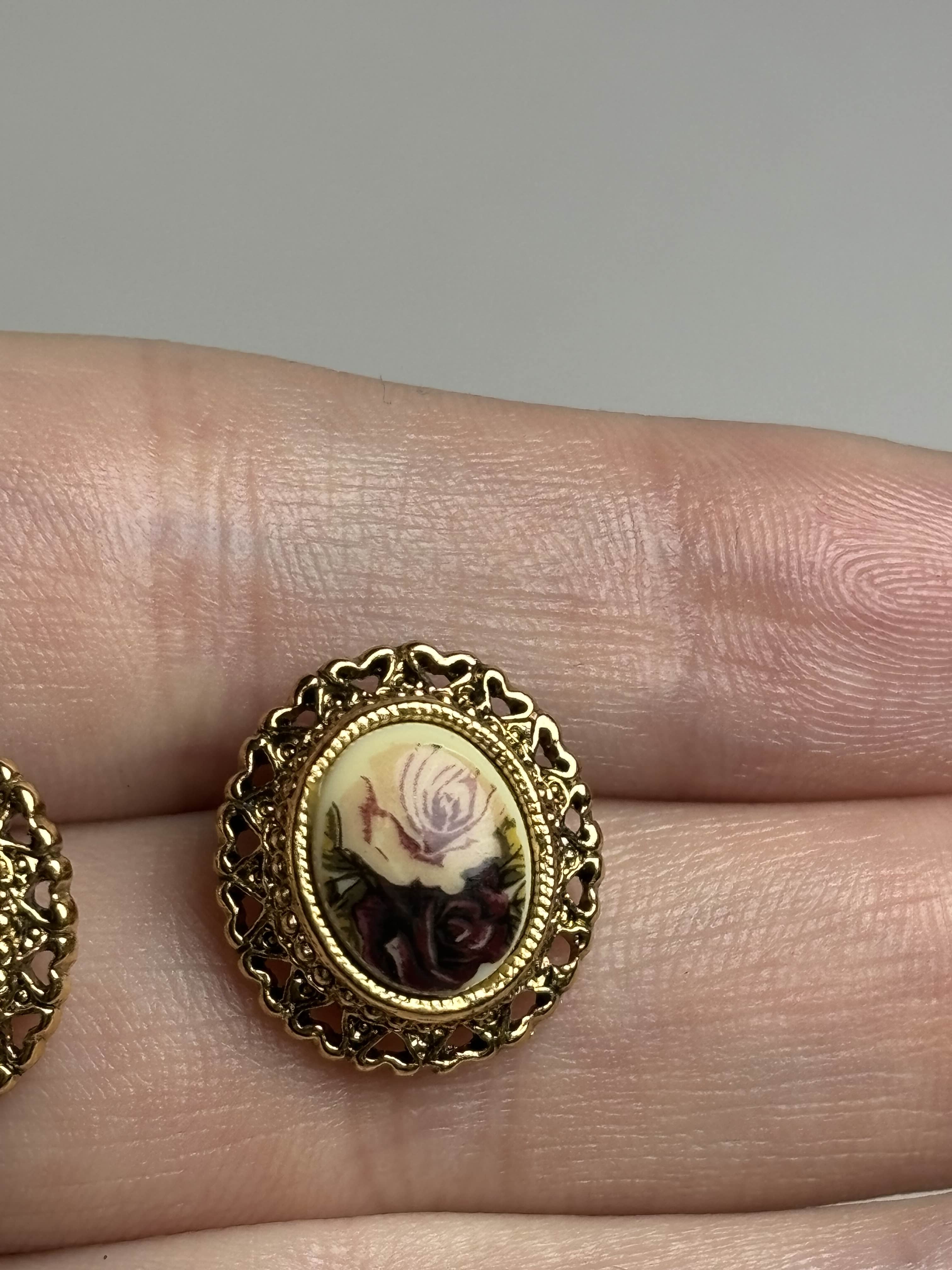 Front of oval earrings with rose design in center and gold filagree heart boarder held in a hand against a neutral background