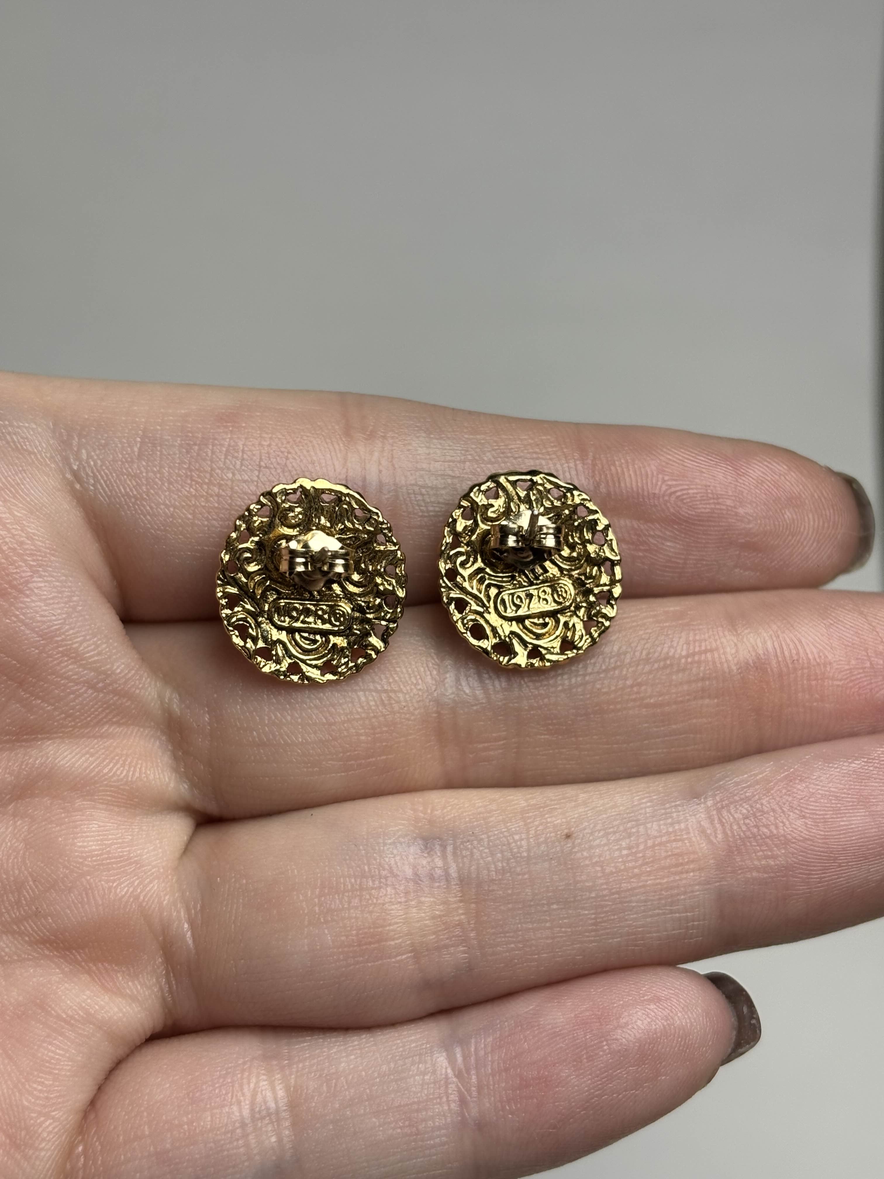 Back of oval earrings with rose design in center and gold filagree heart boarder held in a hand against a neutral background