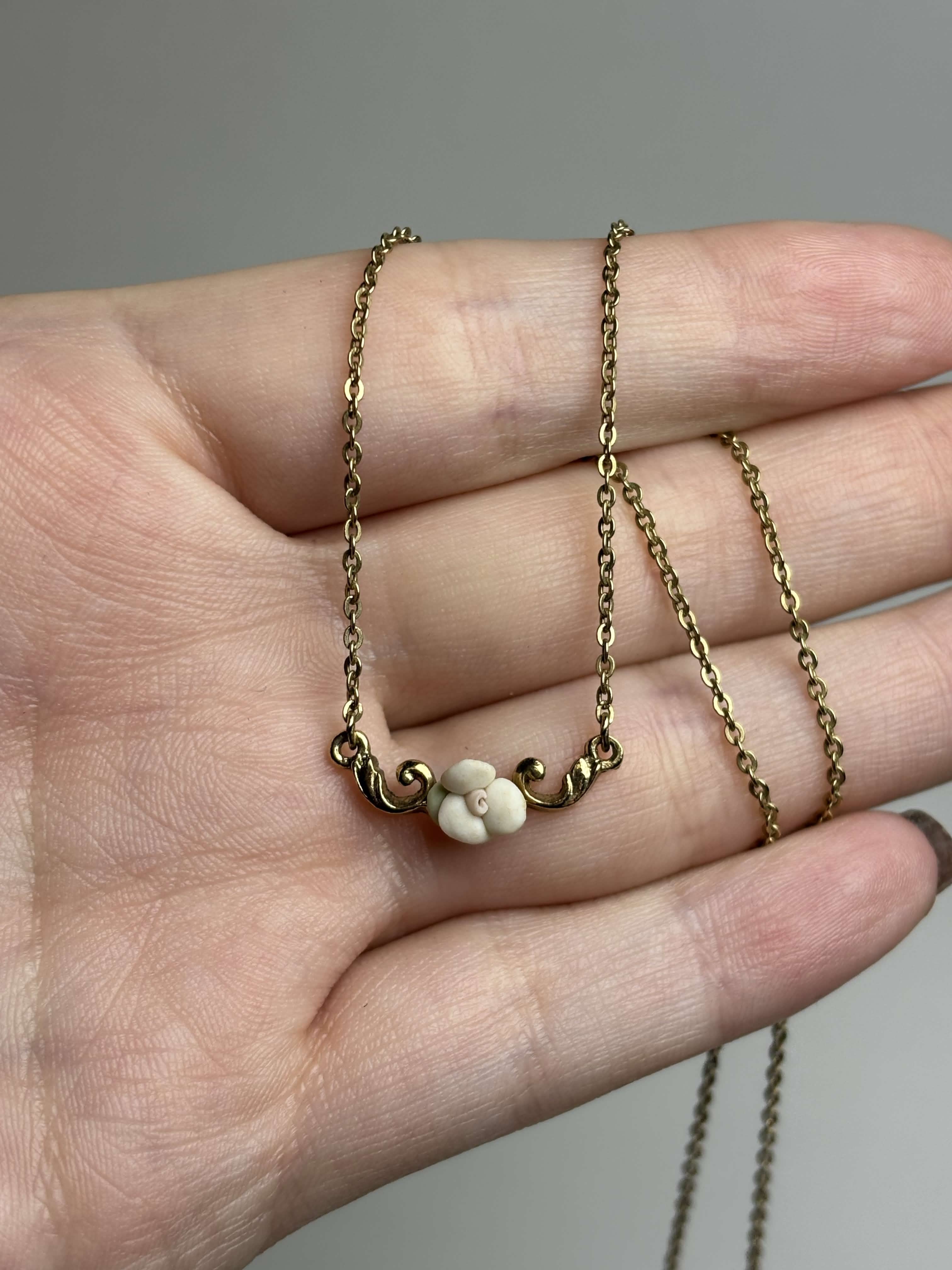 Front of gold necklace with a small white rose pendant held in a hand against a neutral background