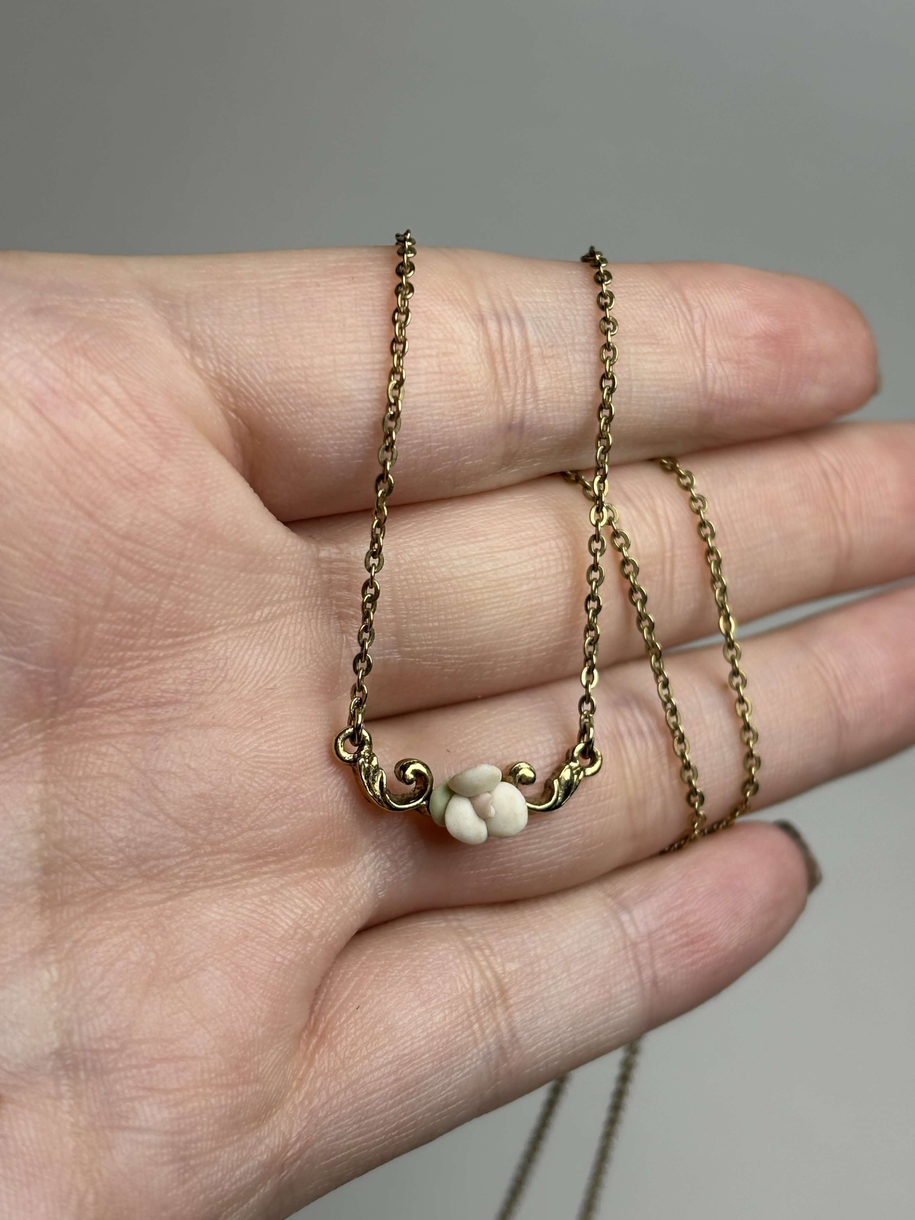 Front of gold necklace with a small white rose pendant held in a hand against a neutral background