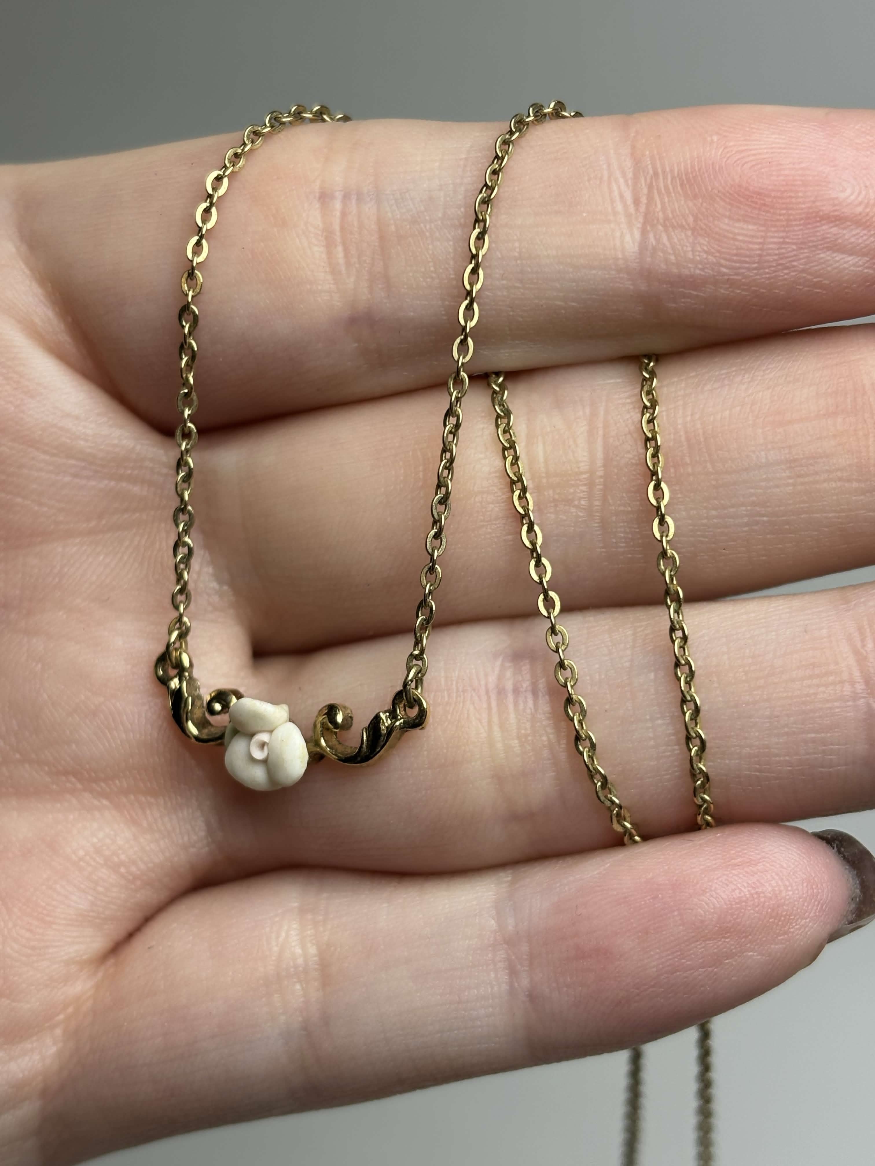 Front of gold necklace with a small white rose pendant held in a hand against a neutral background