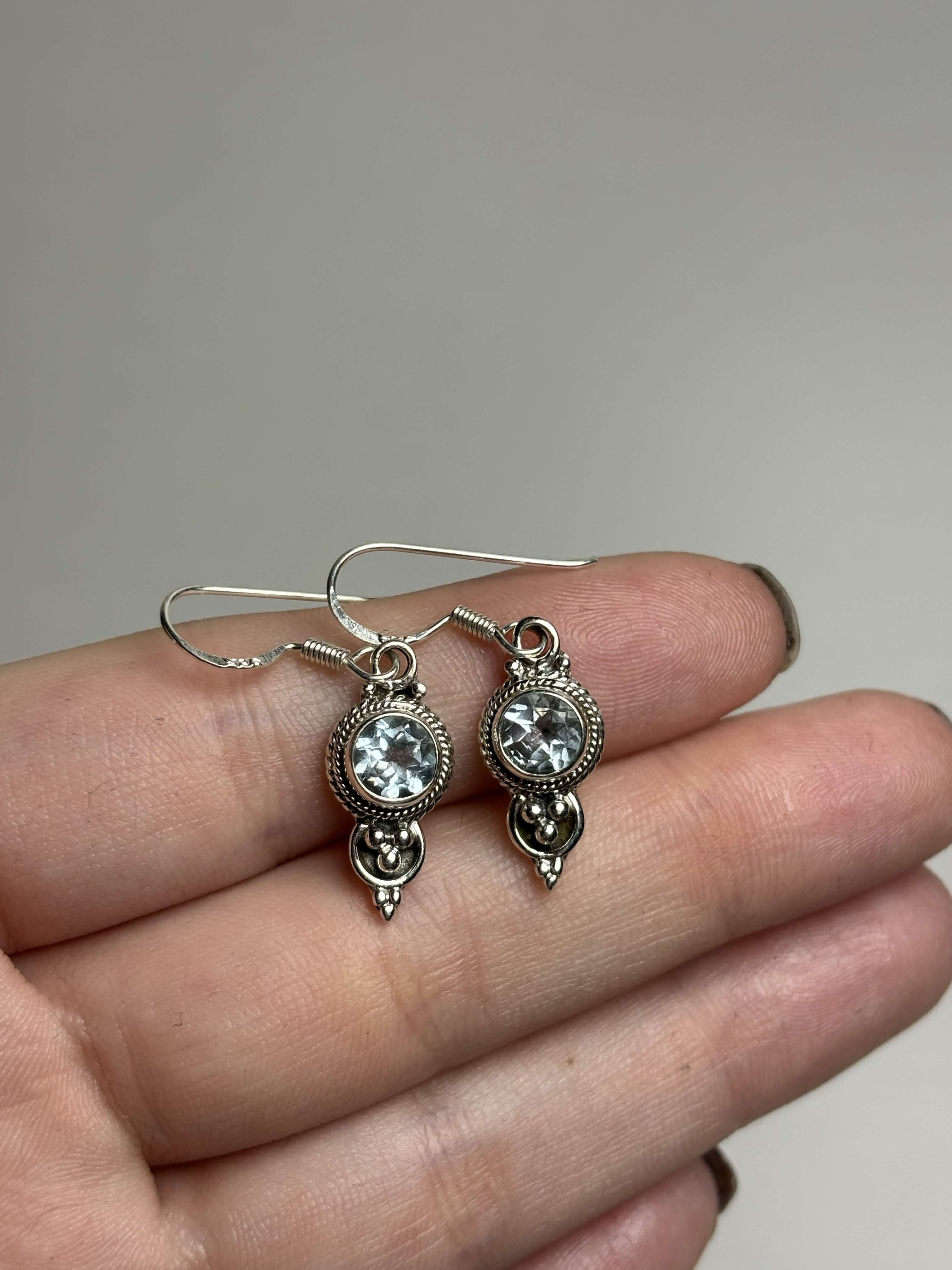 Front of silver dangle earrings with light blue round topaz on a hand with a neutral background