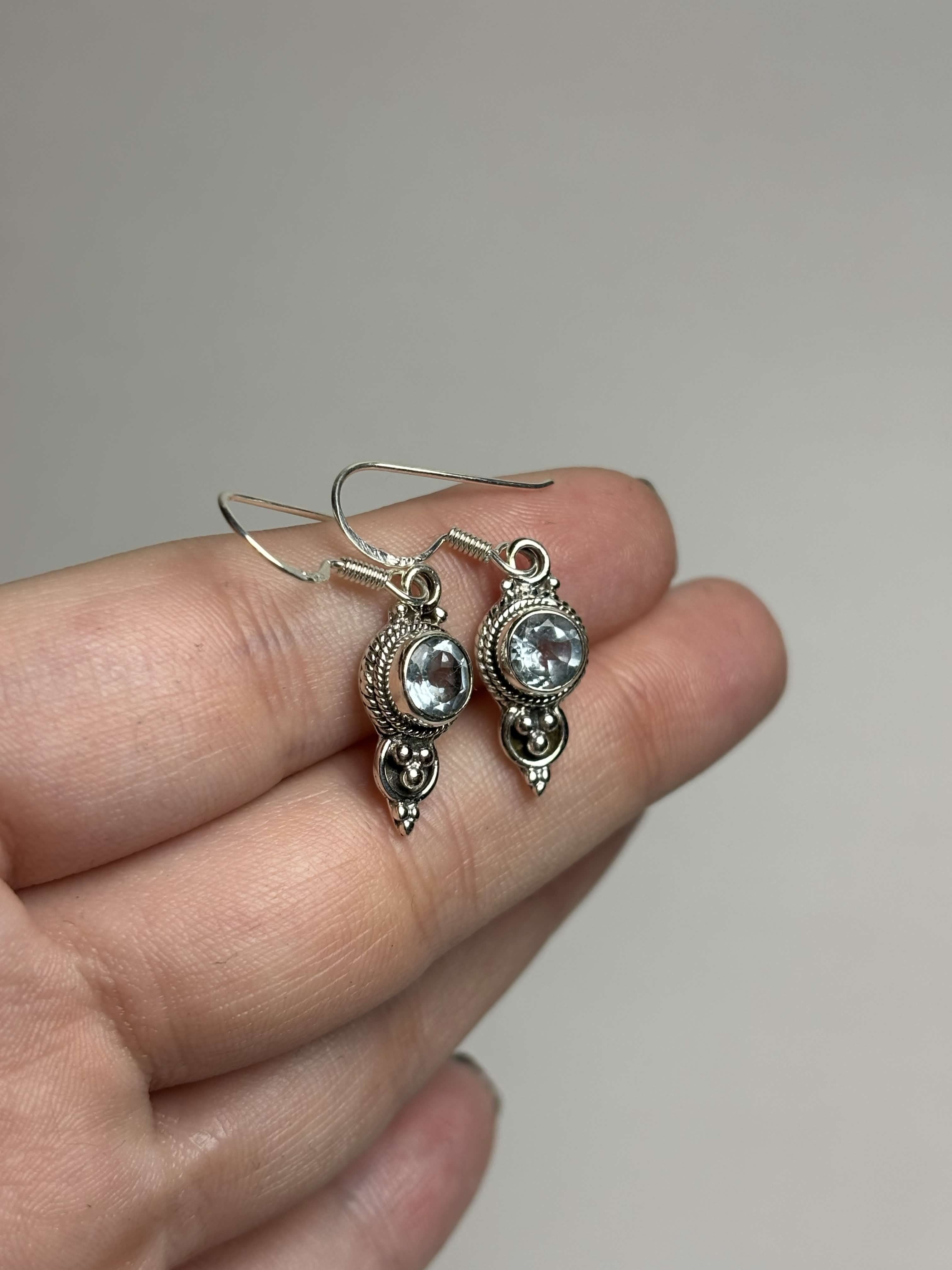 Front of silver dangle earrings with light blue round topaz on a hand with a neutral background