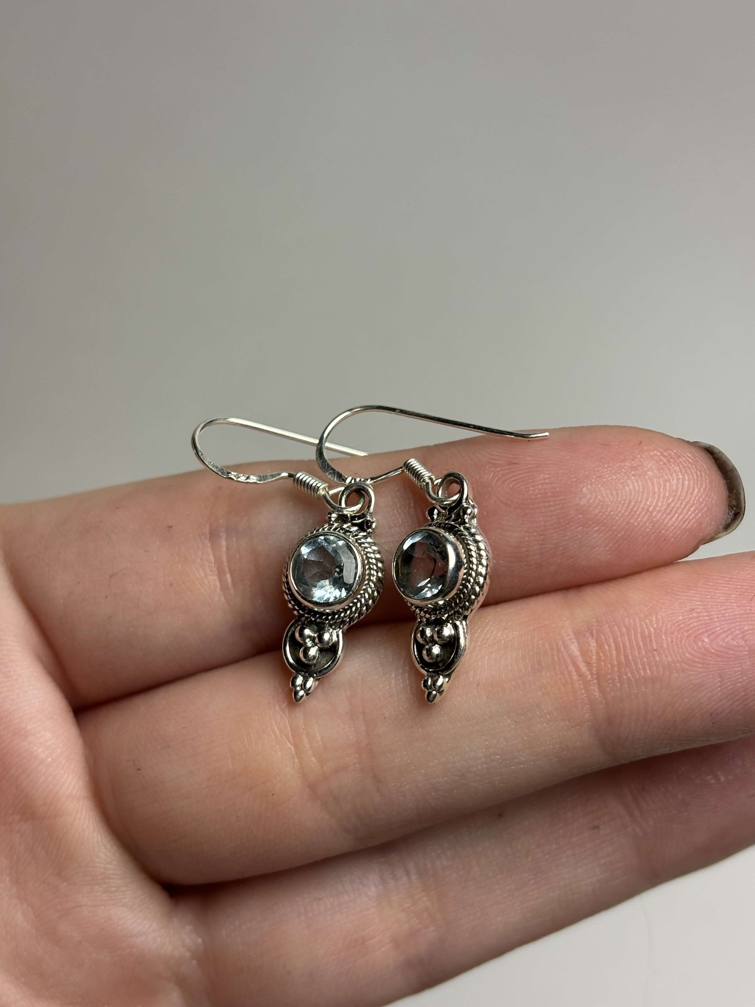 Front of silver dangle earrings with light blue round topaz on a hand with a neutral background