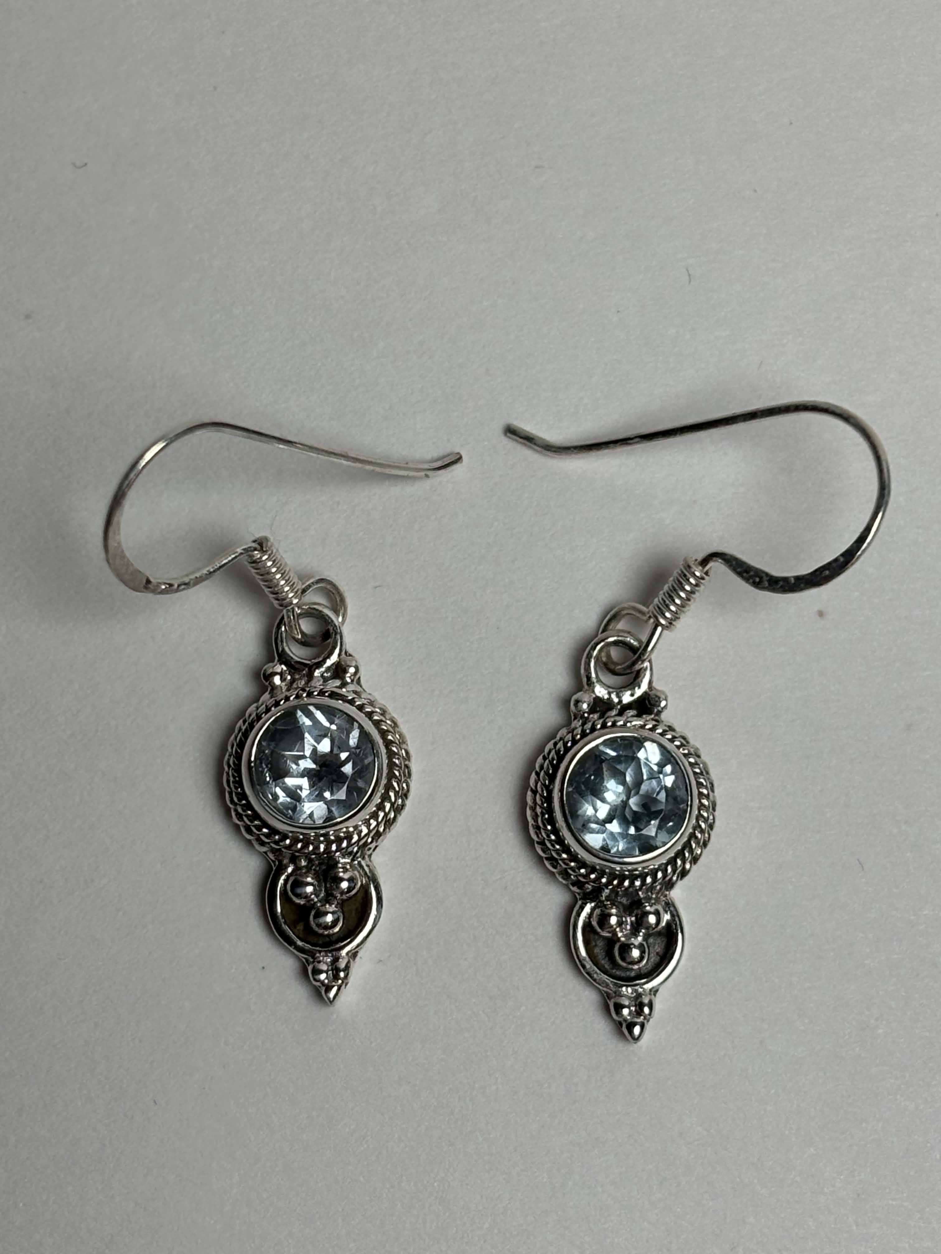 Front of silver dangle earrings with light blue round topaz on a neutral background