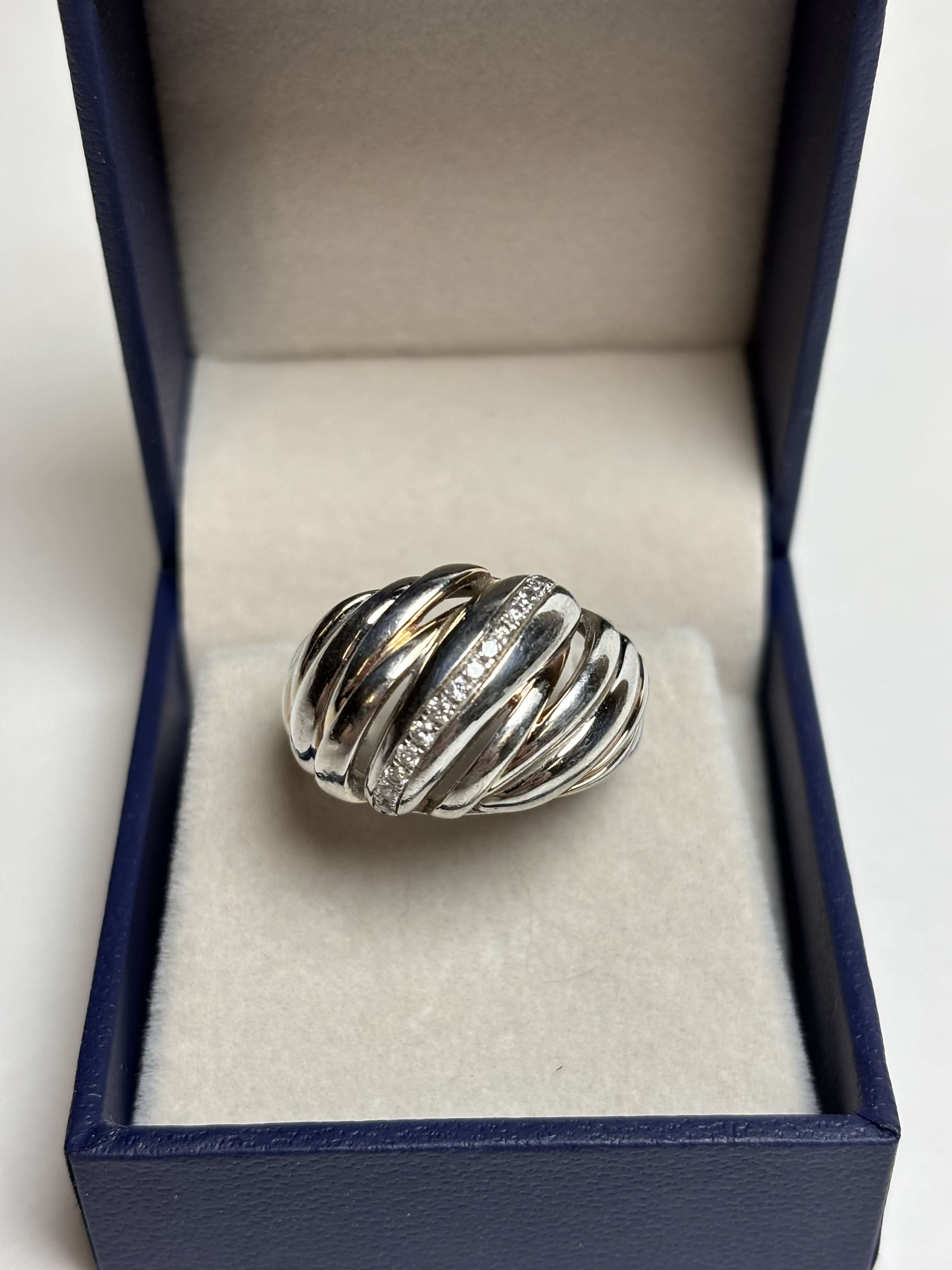 Ribbed / Shrimp / Croissant style silver ring with gold details and a band of pave diamonds in a box on a neutral background
