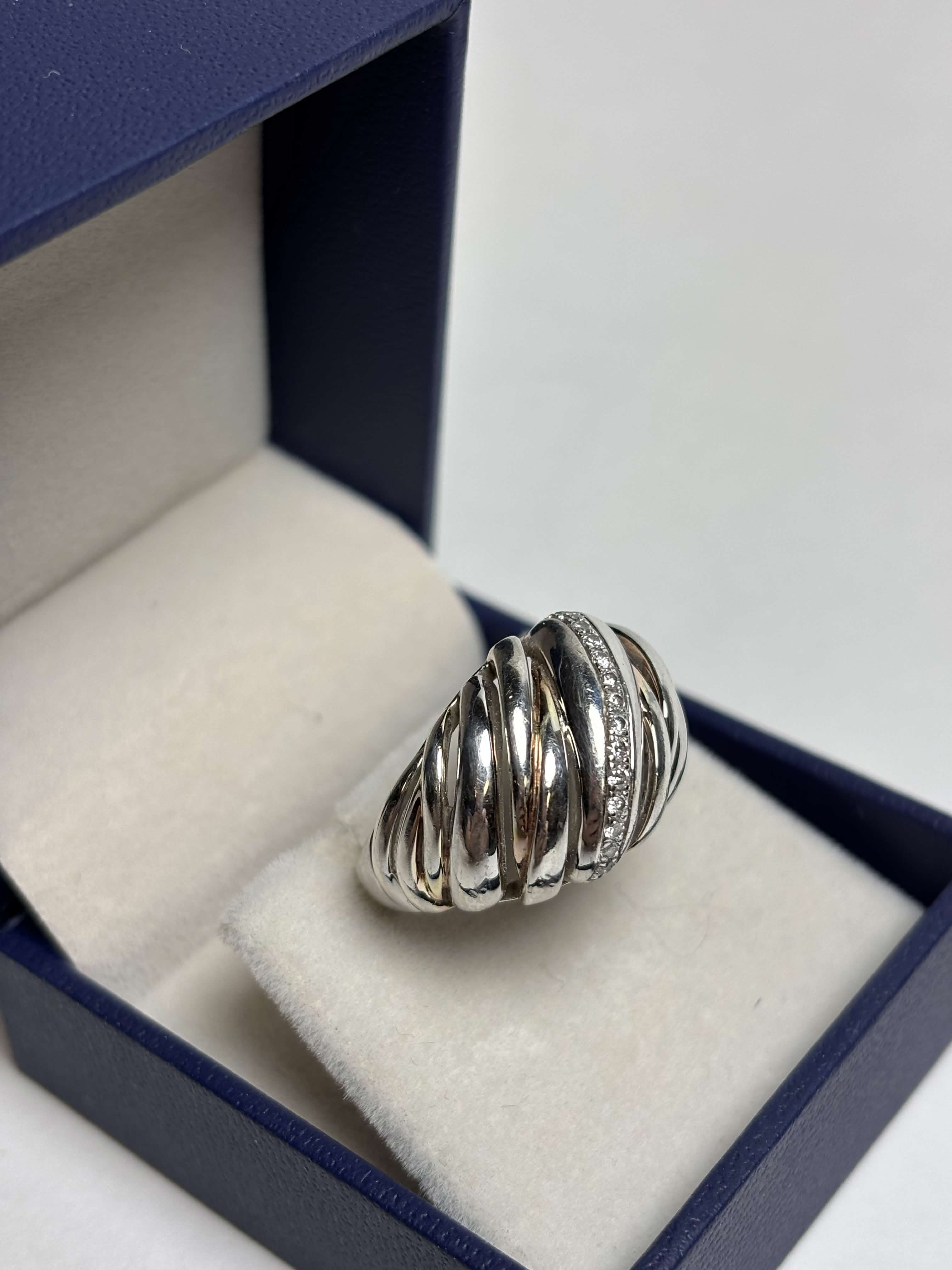 Ribbed / Shrimp / Croissant style silver ring with gold details and a band of pave diamonds in a box on a neutral background