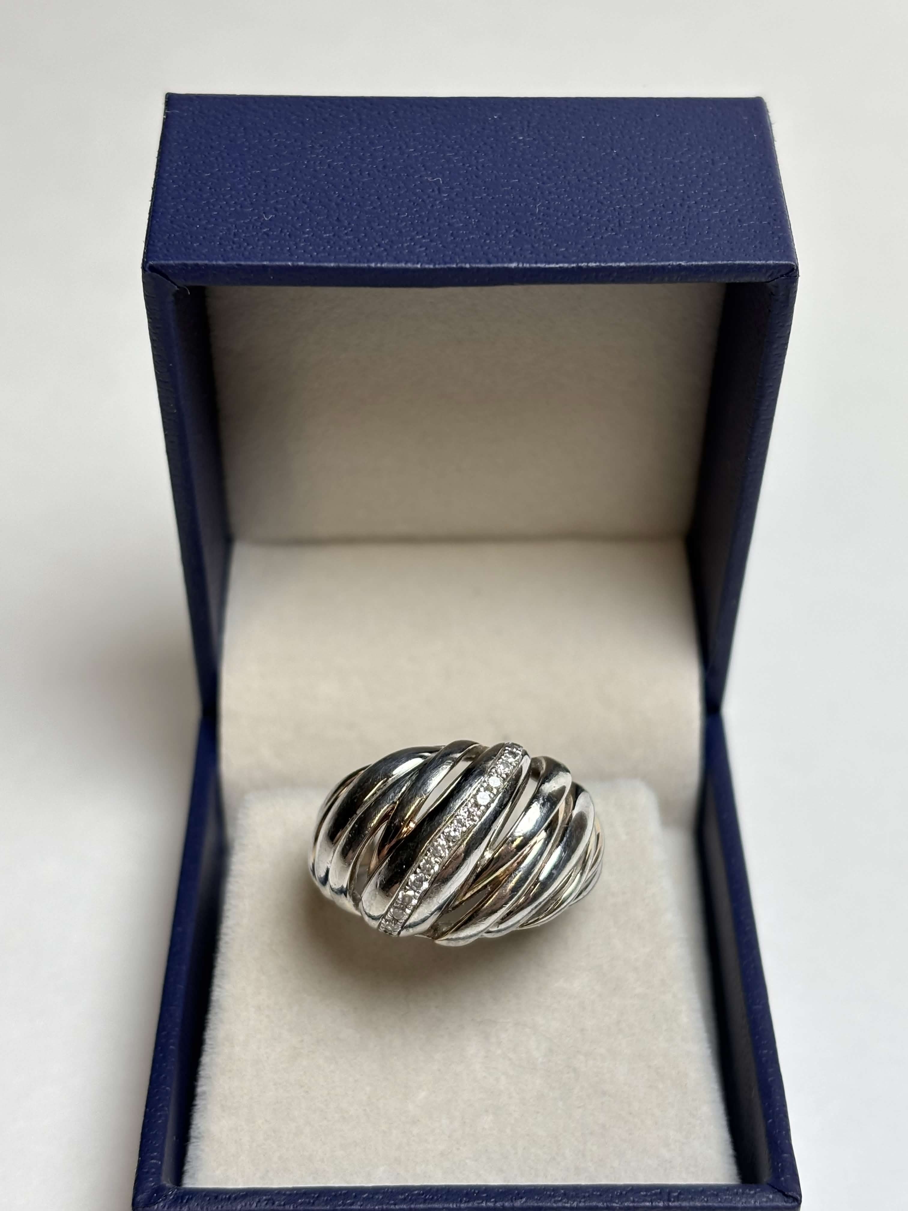 Ribbed / Shrimp / Croissant style silver ring with gold details and a band of pave diamonds in a box on a neutral background