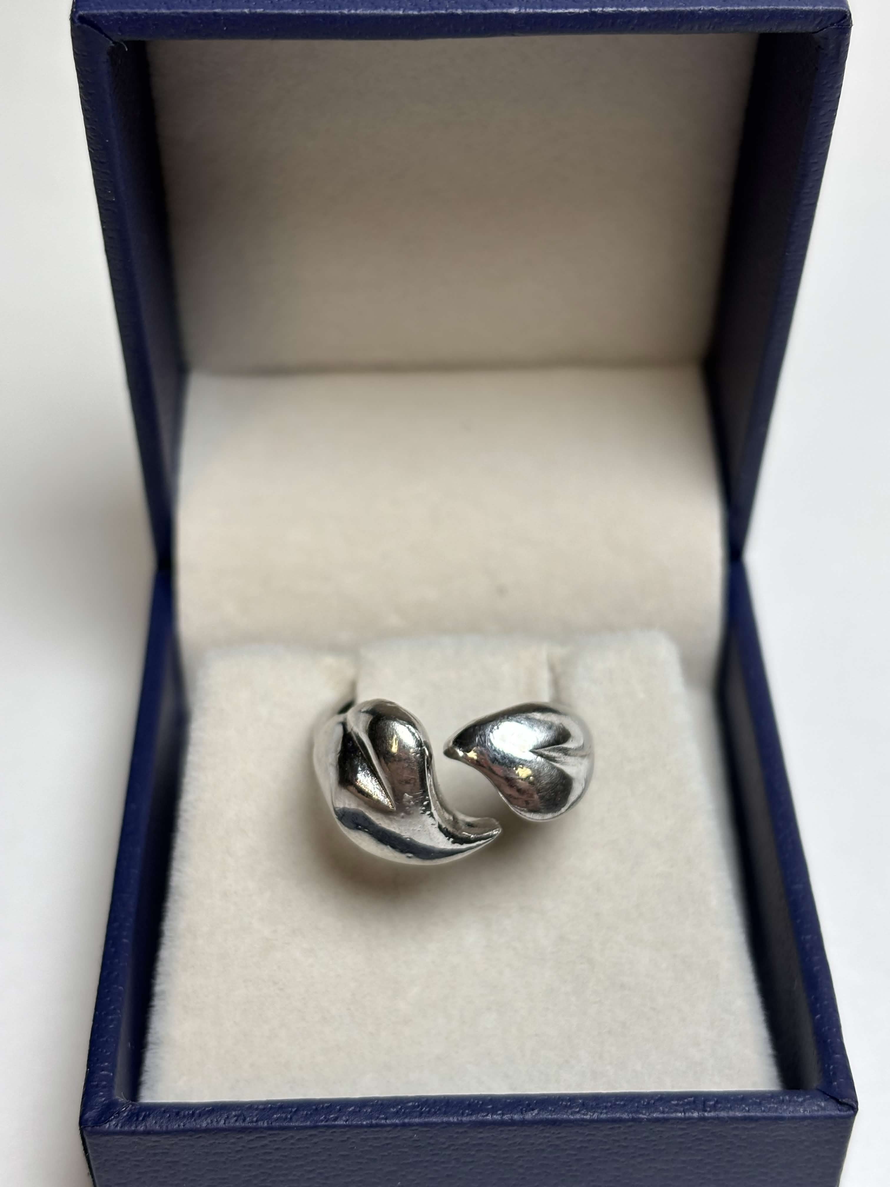 Front of silver tone ring with two heart shaped bypass in a box on a neutral background