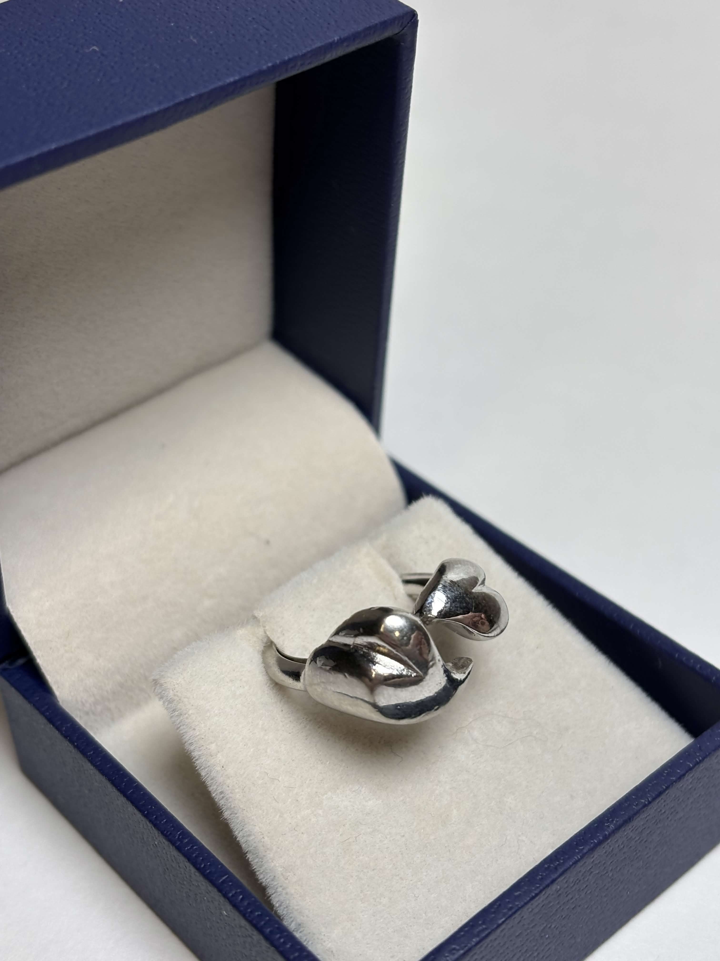 Front of silver tone ring with two heart shaped bypass in a box on a neutral background