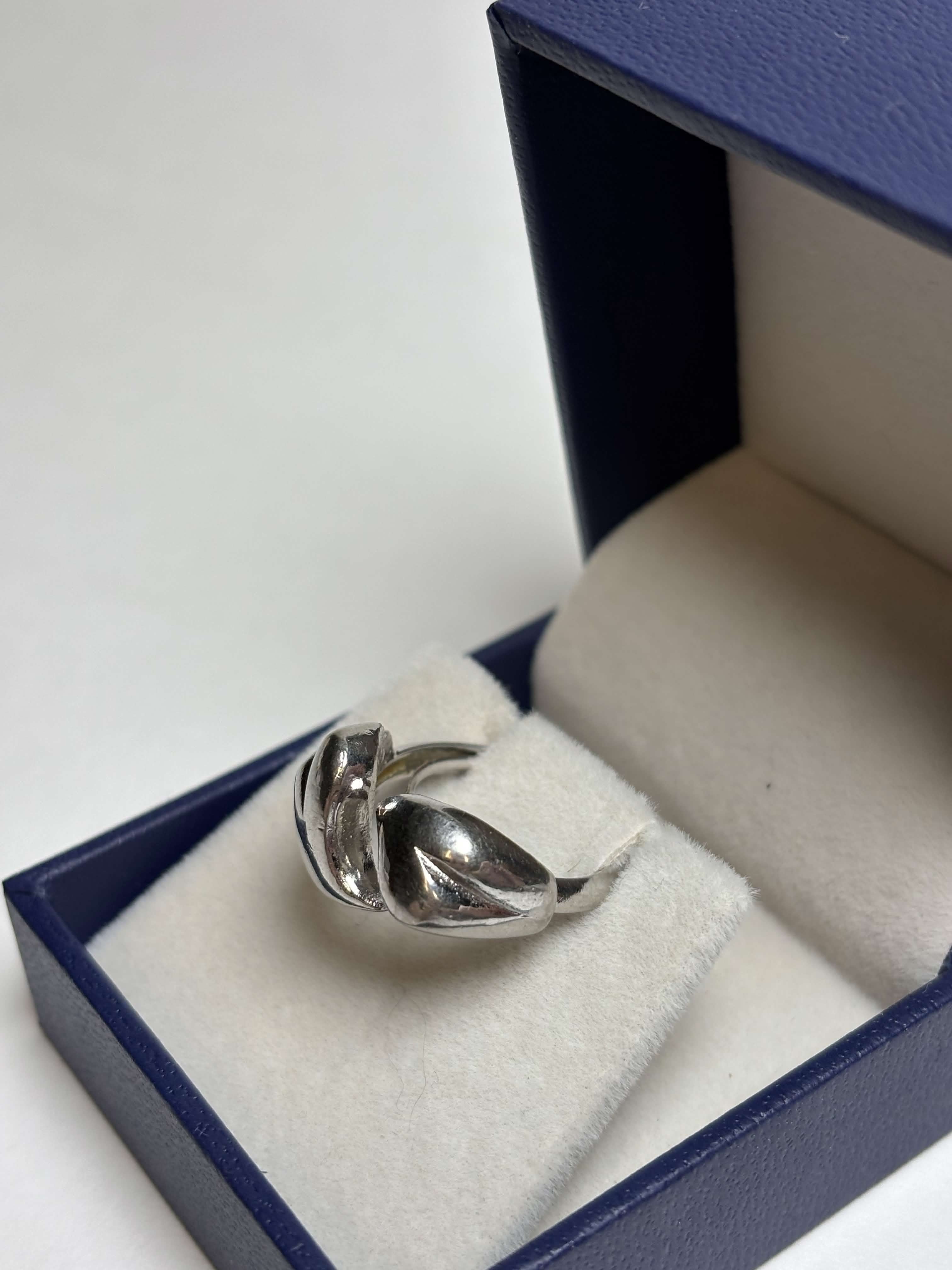 Front of silver tone ring with two heart shaped bypass in a box on a neutral background