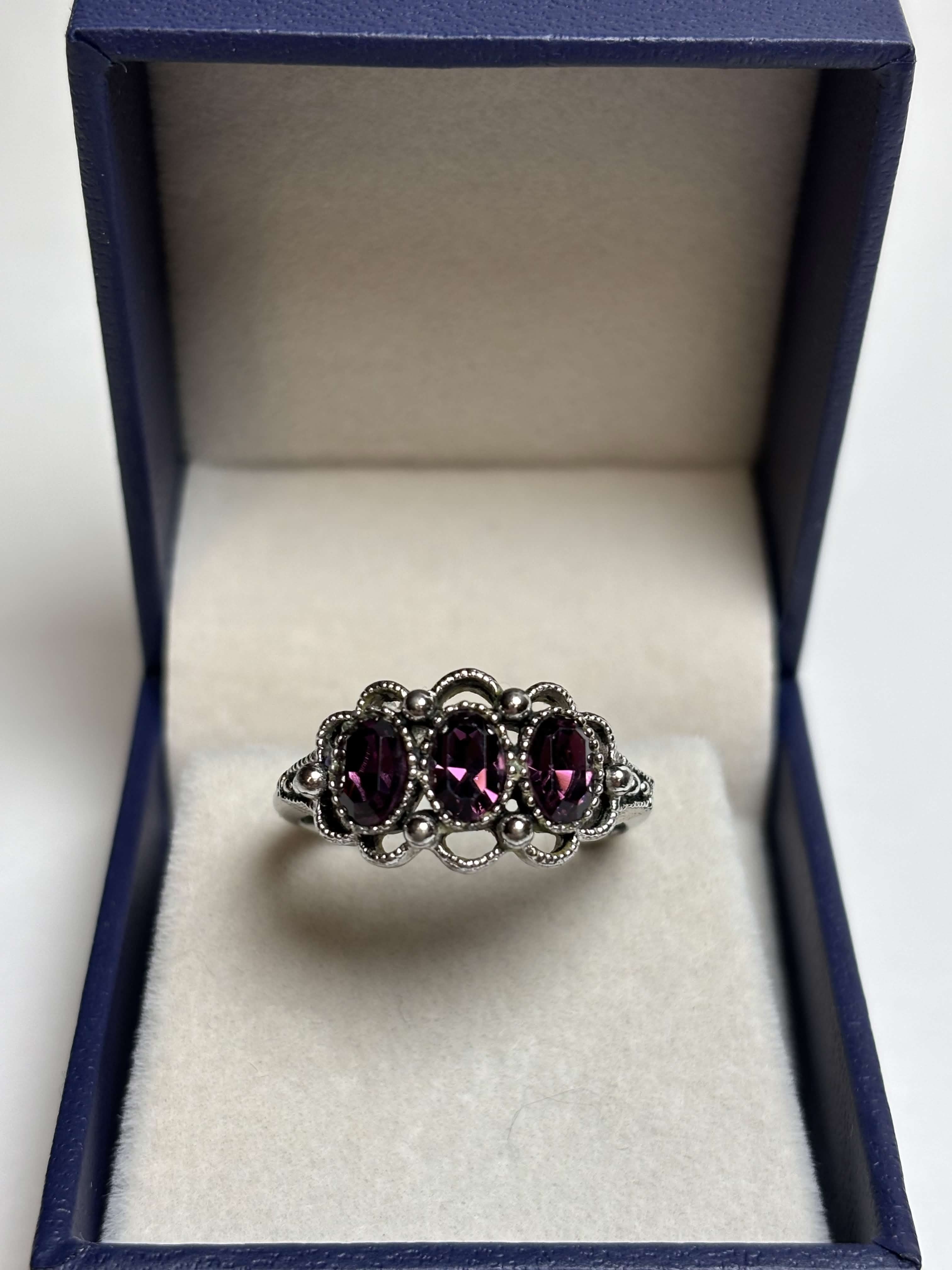 Front of silver ring with three purple stones in a box on a neutral background