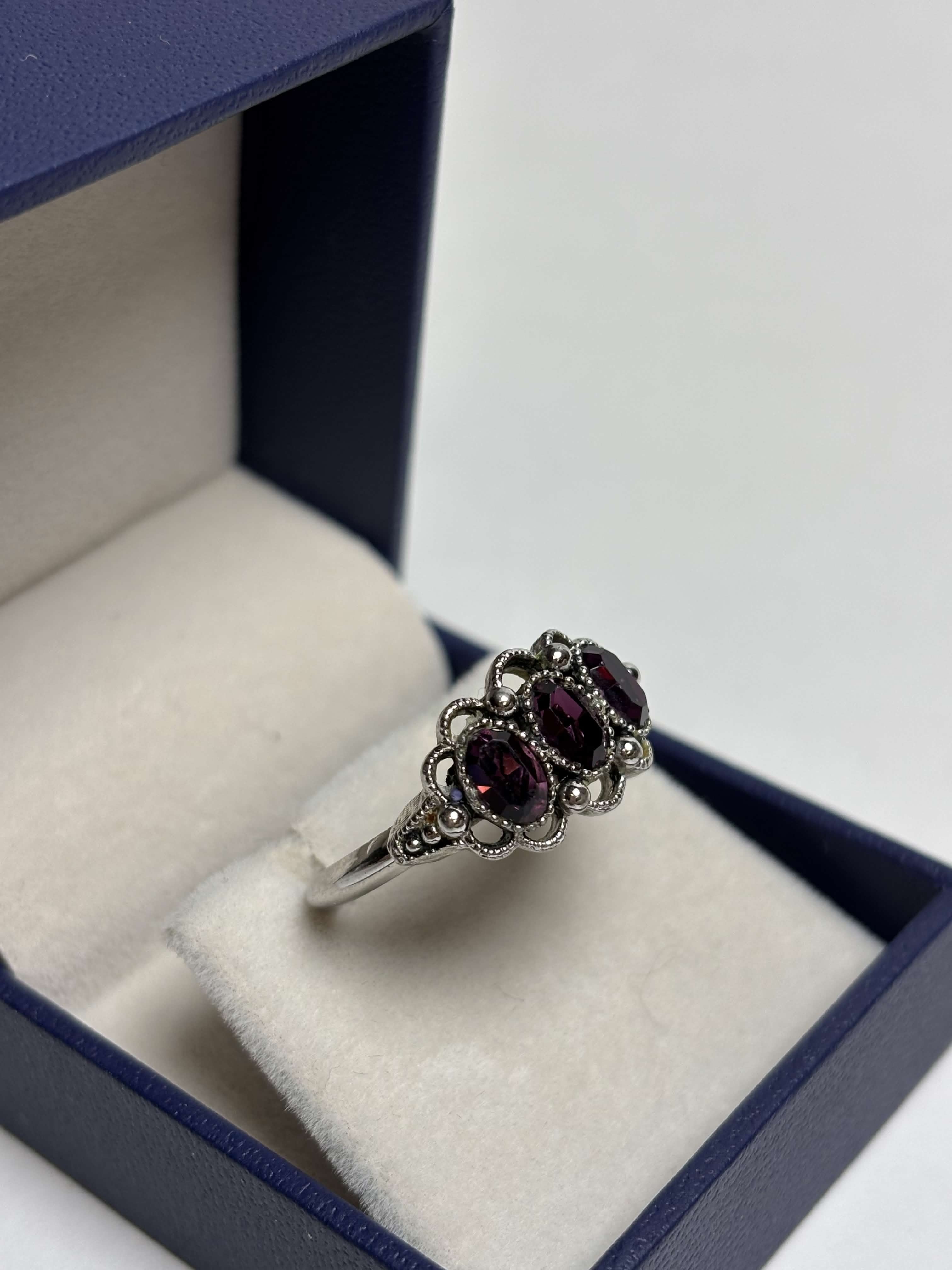 Front of silver ring with three purple stones in a box on a neutral background