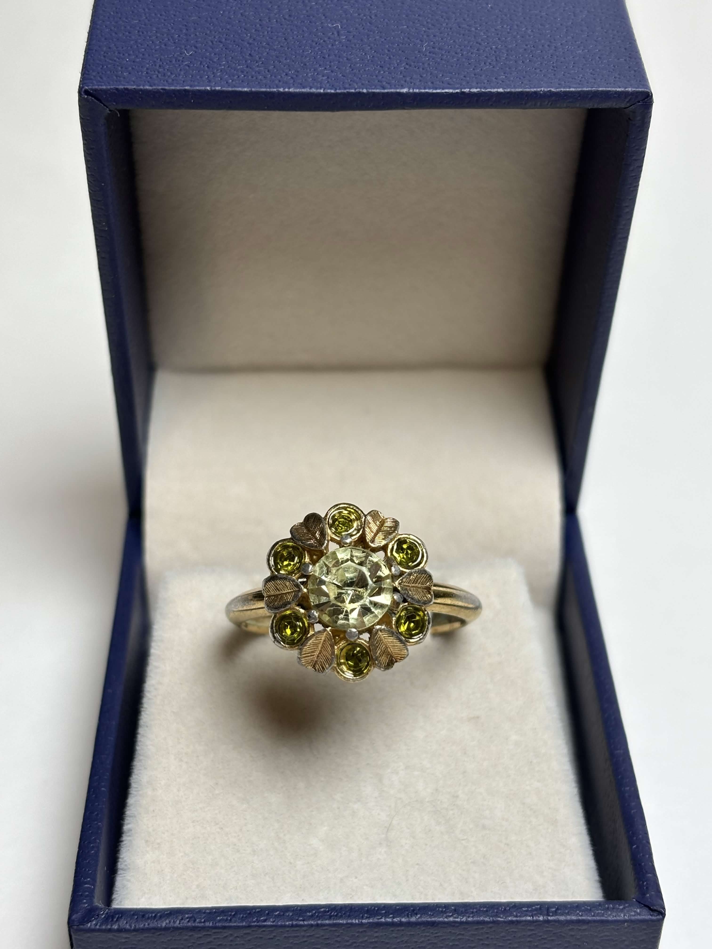 Front of gold ring with light green center stone and alternating gold leaves and darker green round stones ring in a box on a neutral background