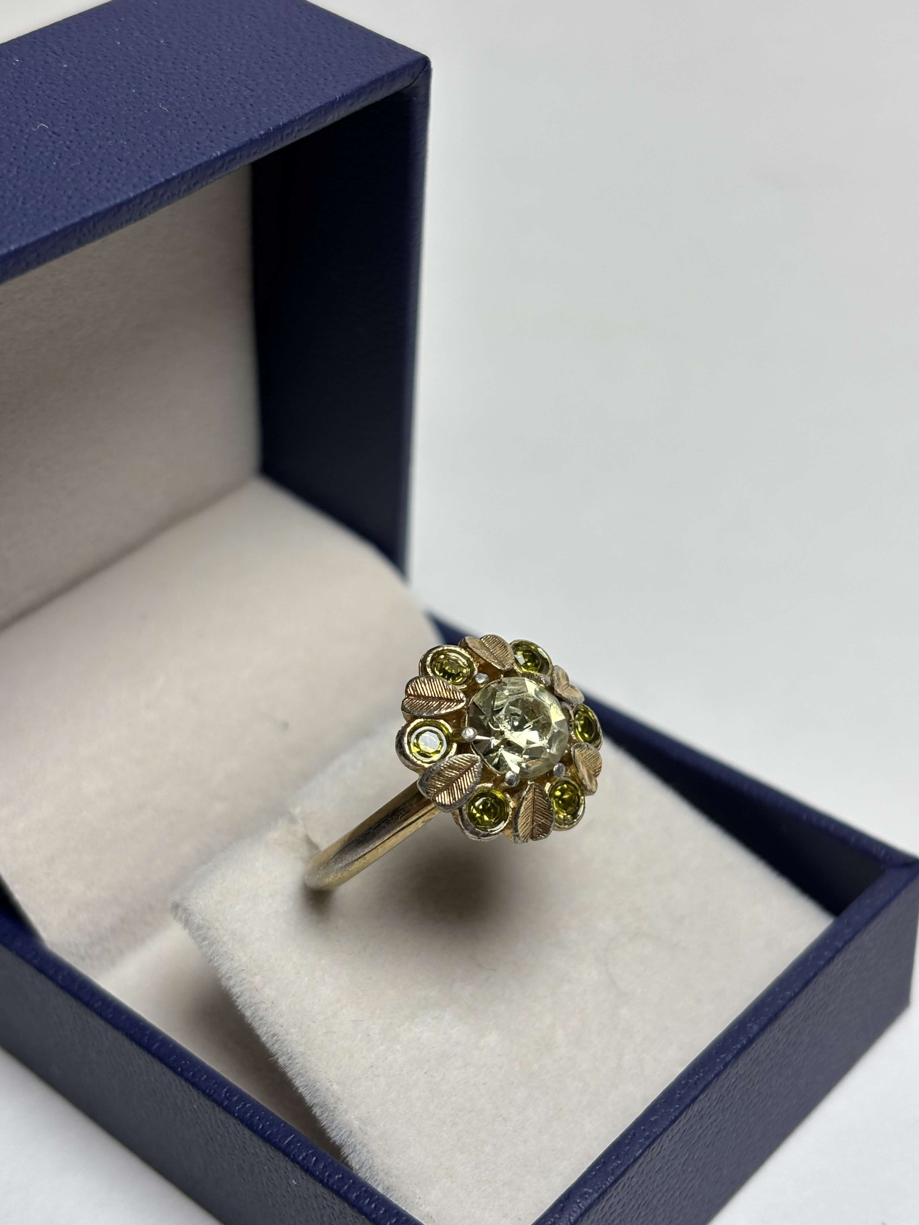Front of gold ring with light green center stone and alternating gold leaves and darker green round stones ring in a box on a neutral background