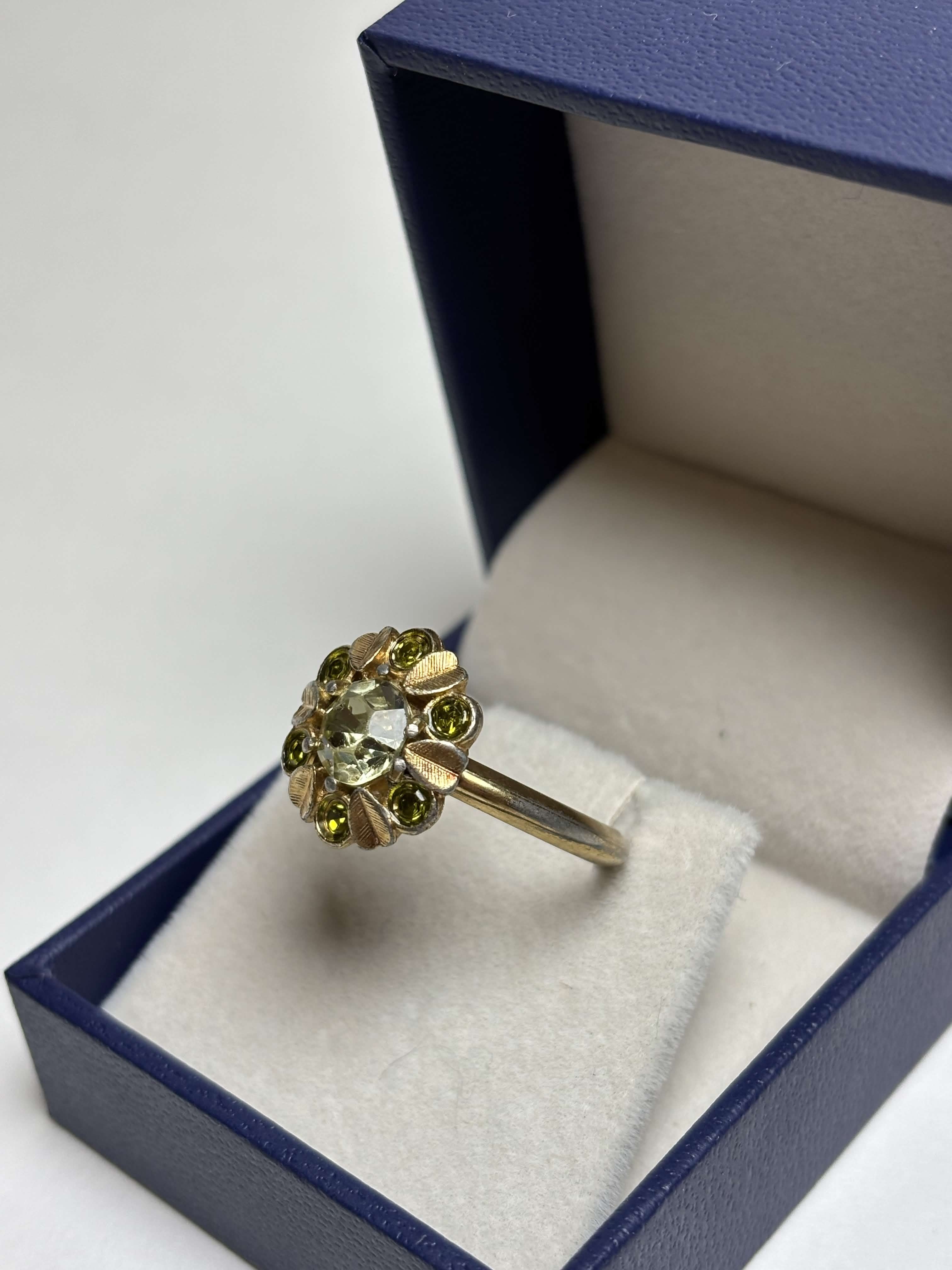 Front of gold ring with light green center stone and alternating gold leaves and darker green round stones ring in a box on a neutral background