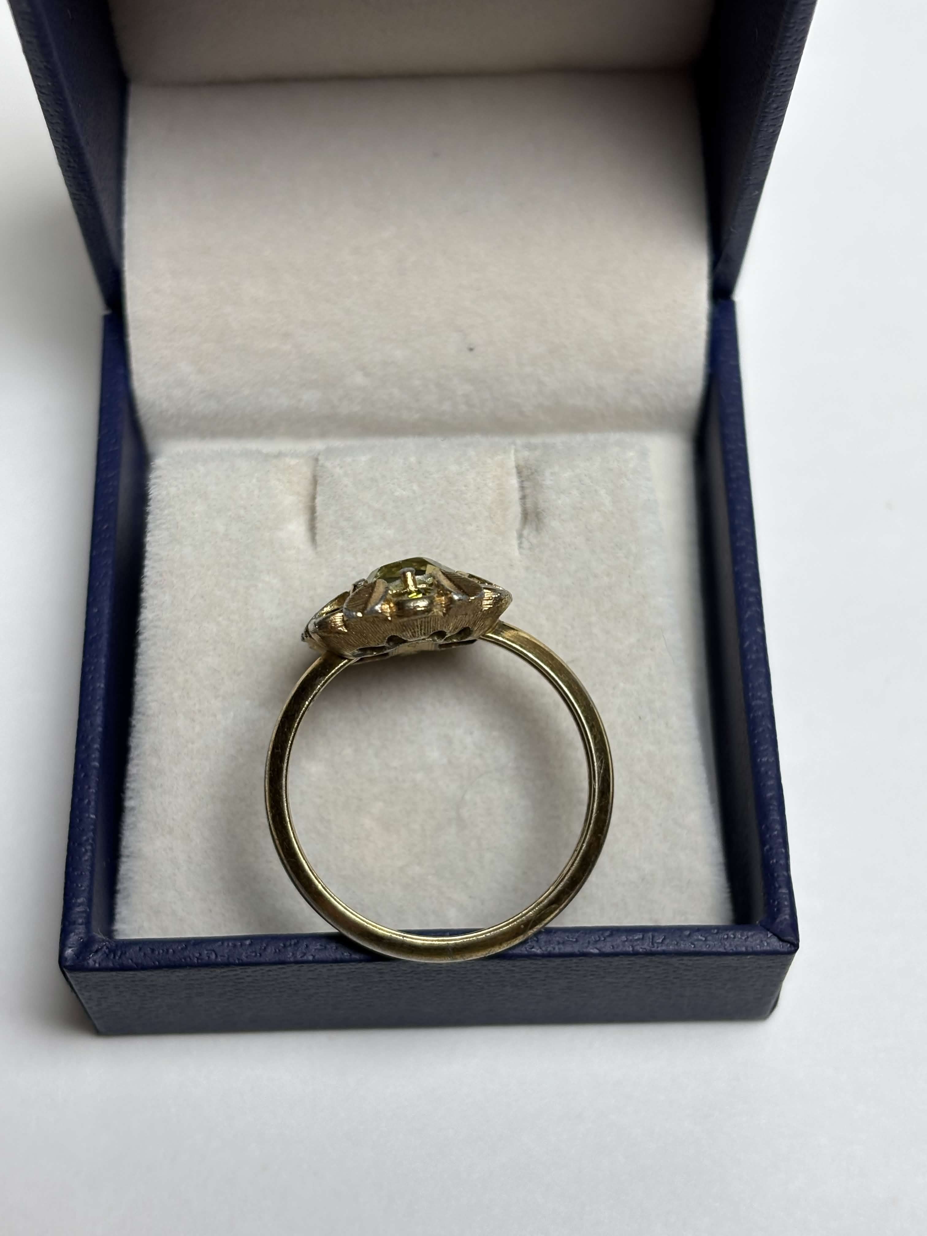 Front of gold ring with light green center stone and alternating gold leaves and darker green round stones ring in a box on a neutral background