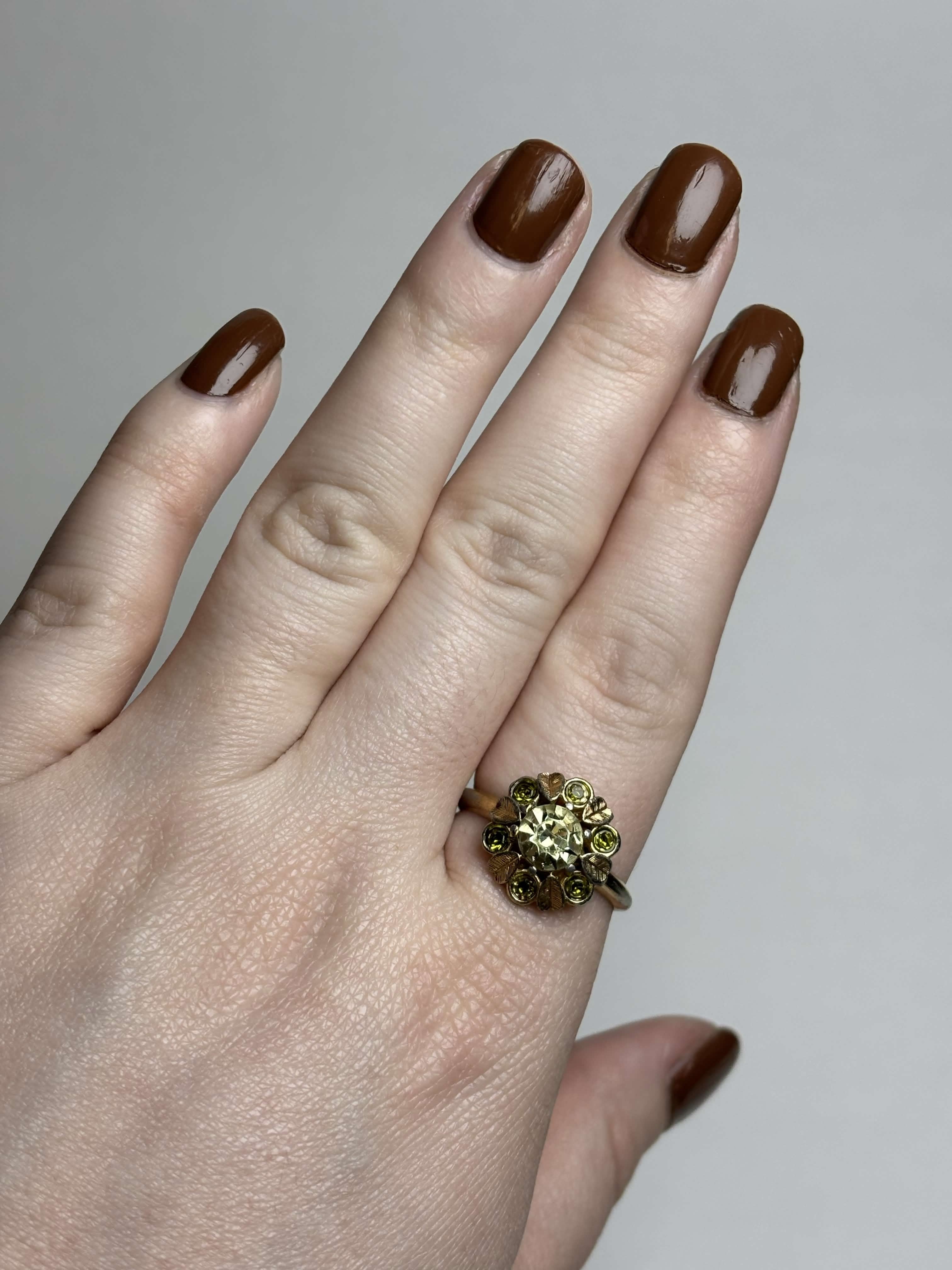 Front of gold ring with light green center stone and alternating gold leaves and darker green round stones ring on a hand on a neutral background