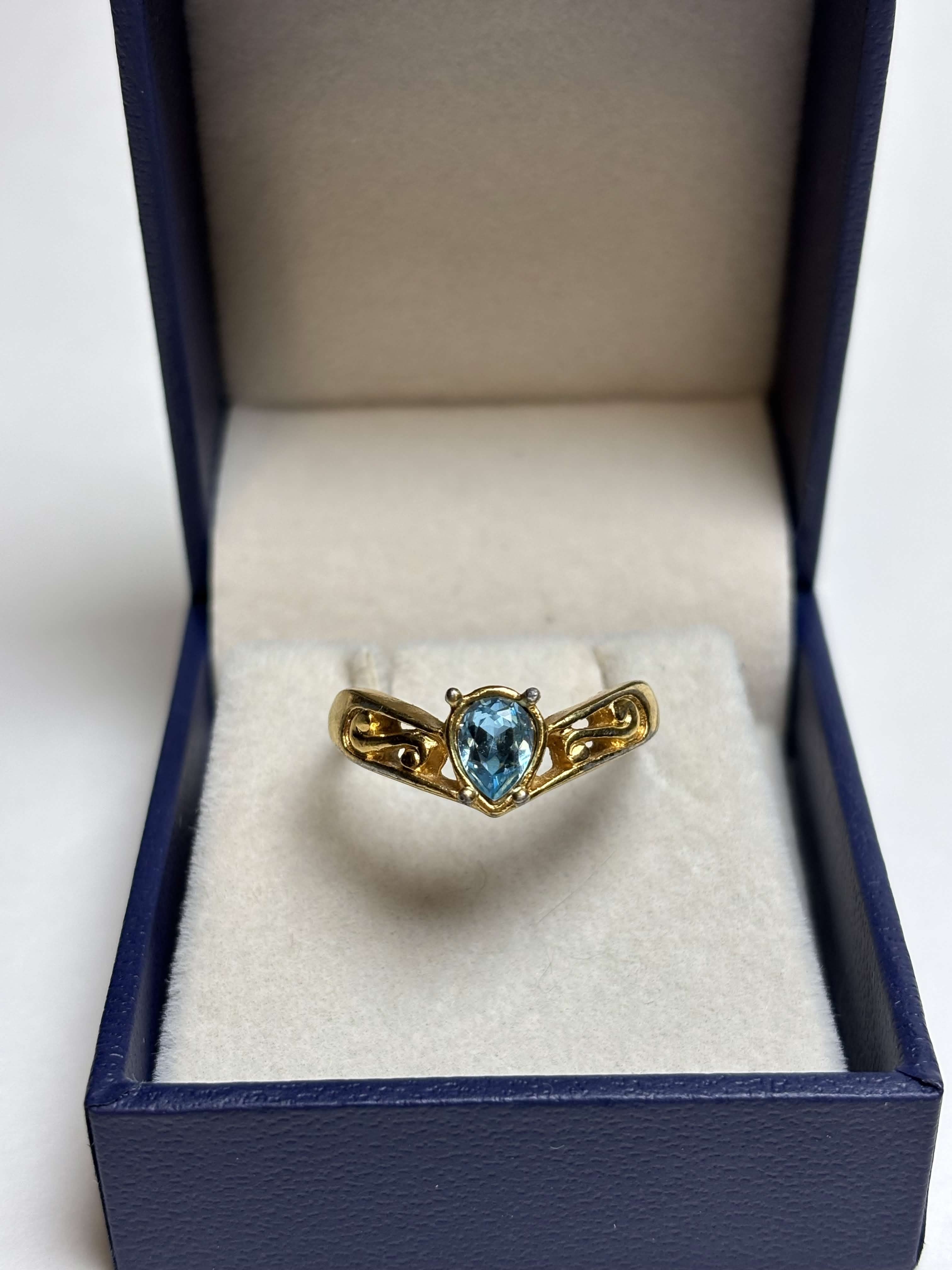 Front of gold tone ring with a pear shaped blue stone in the middle and filigree gold design on the sides in a box on a neutral background