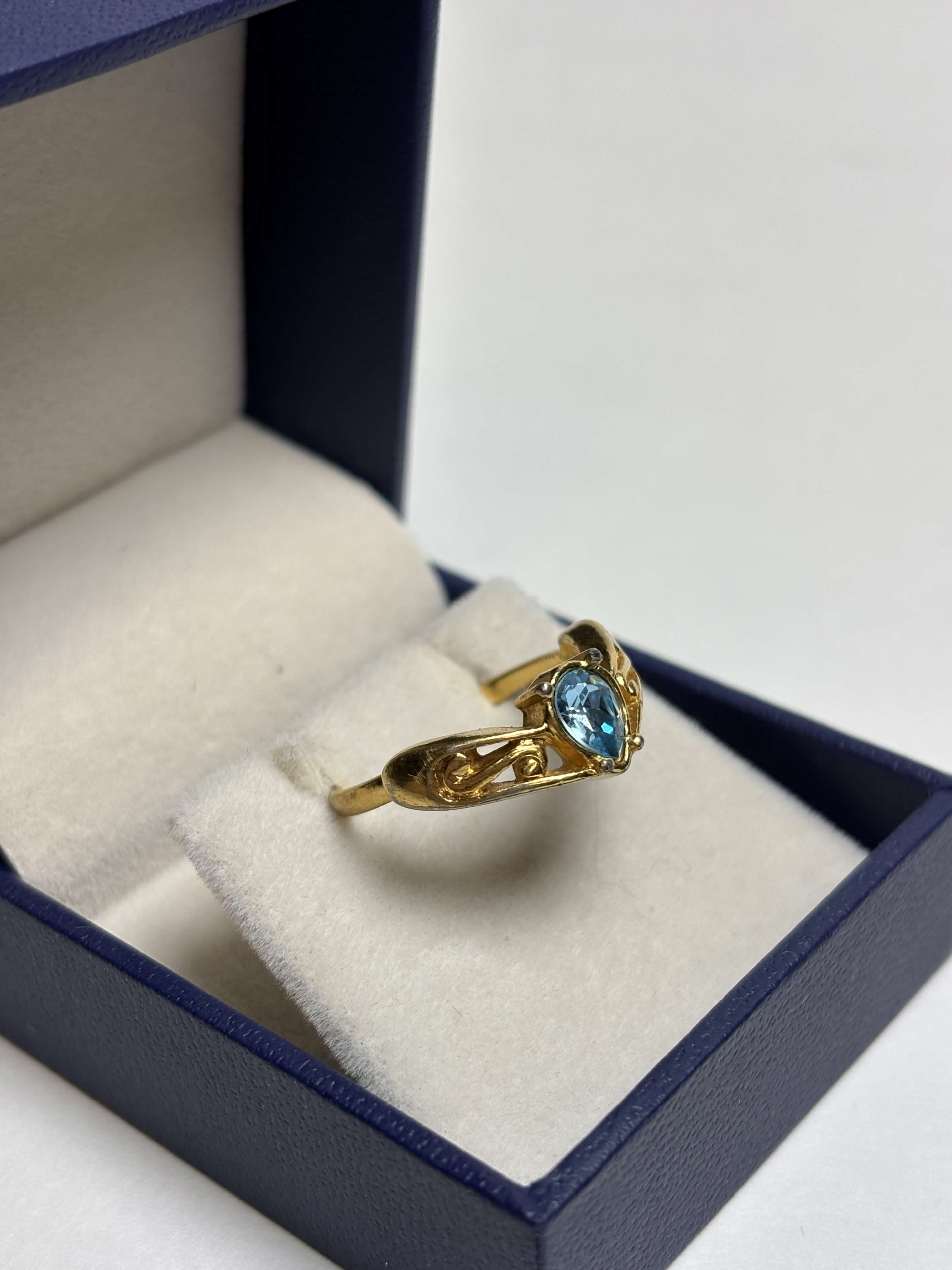 Front of gold tone ring with a pear shaped blue stone in the middle and filigree gold design on the sides in a box on a neutral background