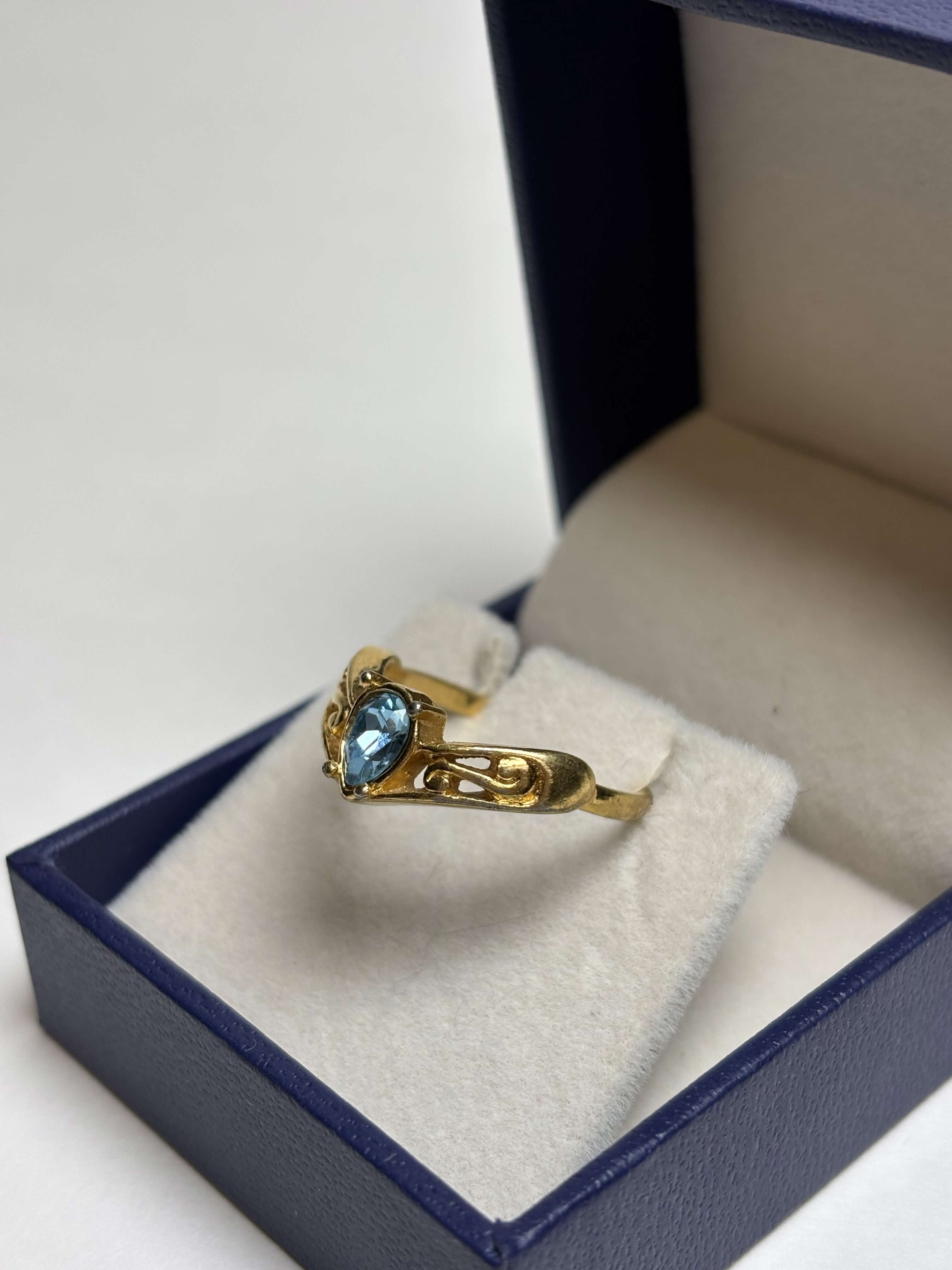 Front of gold tone ring with a pear shaped blue stone in the middle and filigree gold design on the sides in a box on a neutral background