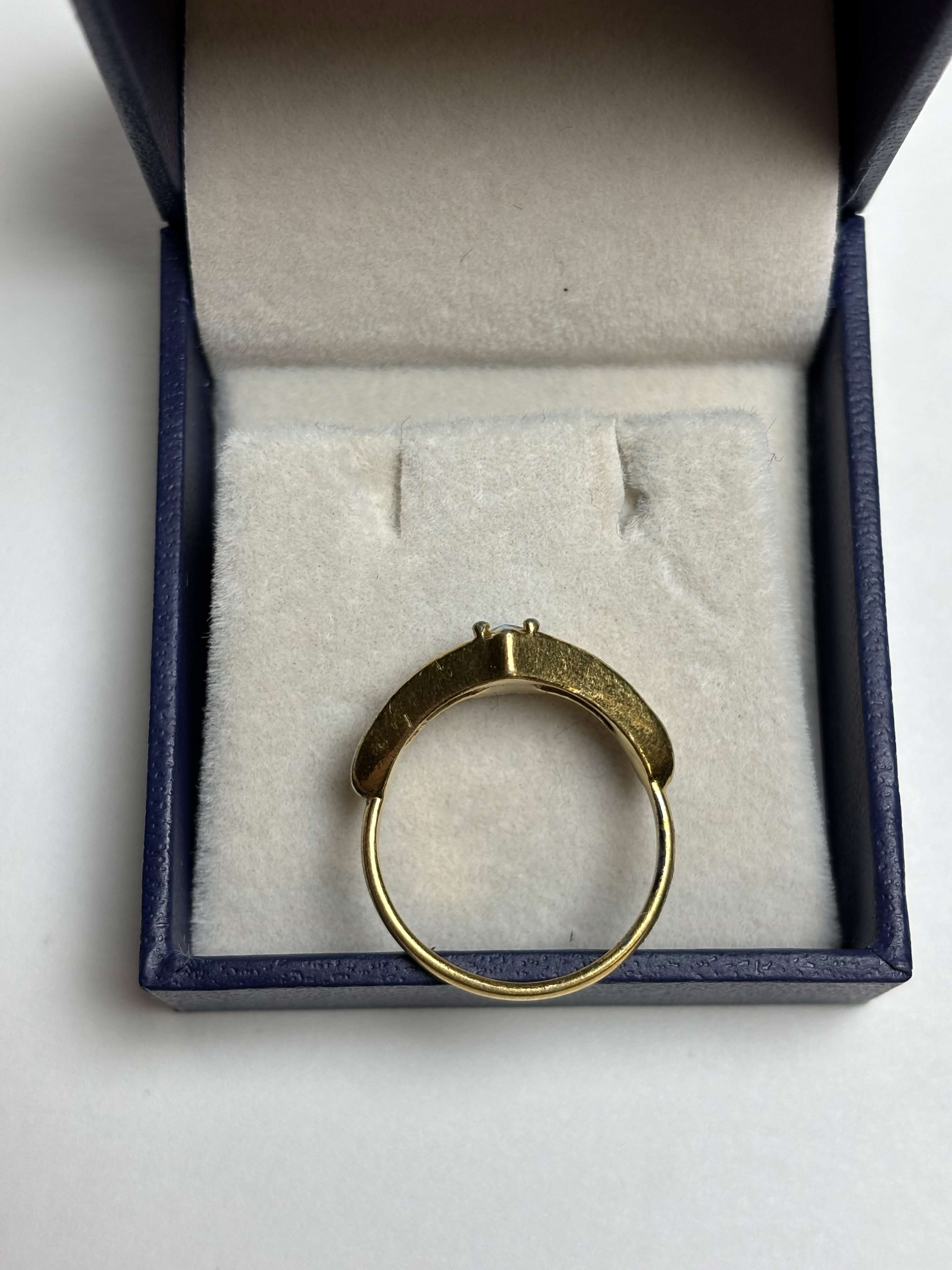 Side of gold tone ring with a pear shaped blue stone in the middle and filigree gold design on the sides in a box on a neutral background