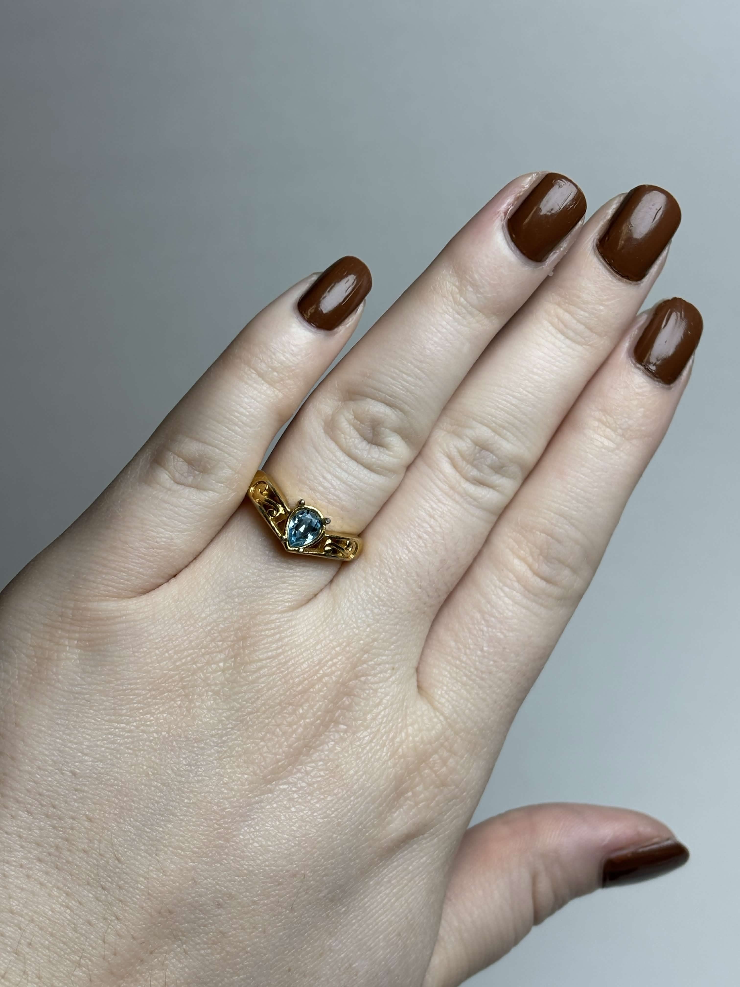 Front of gold tone ring with a pear shaped blue stone in the middle and filigree gold design on the sides on a hand on a neutral background