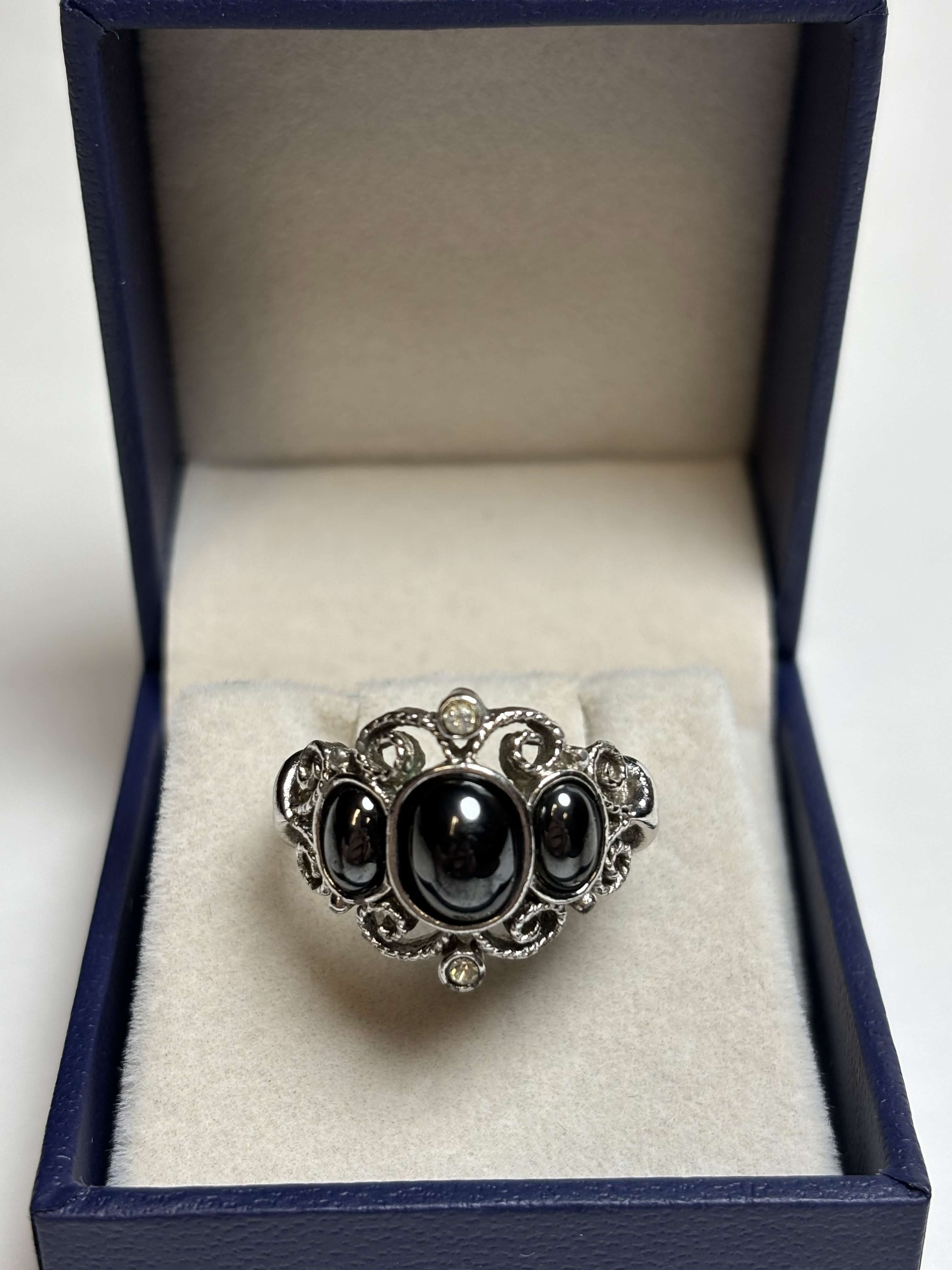 Front of silver tone ring with three oval black stones in the middle surrounded by filagree and has two clear stones at the highest points in a box on a neutral background