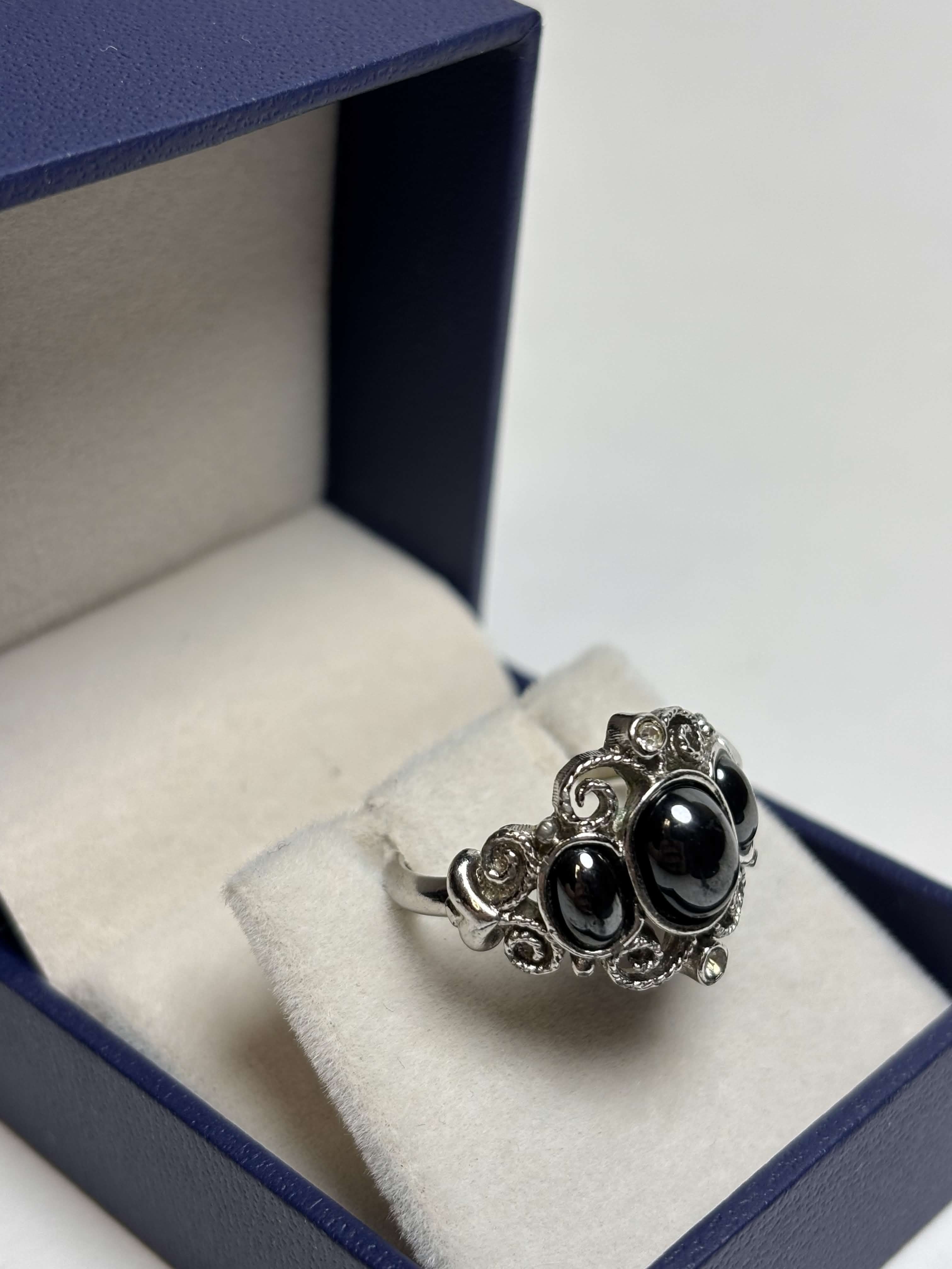 Front of silver tone ring with three oval black stones in the middle surrounded by filagree and has two clear stones at the highest points in a box on a neutral background