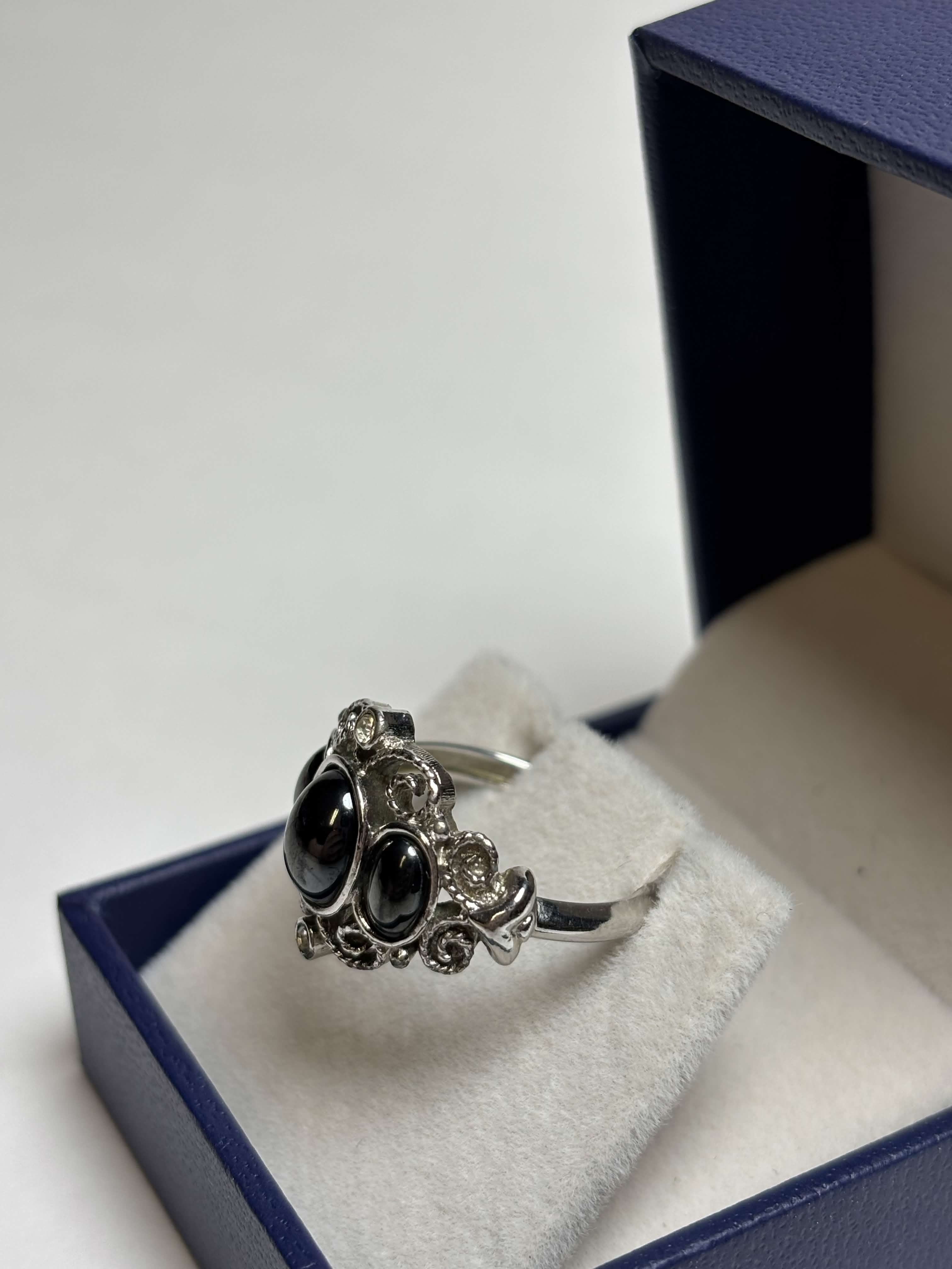 Front of silver tone ring with three oval black stones in the middle surrounded by filagree and has two clear stones at the highest points in a box on a neutral background