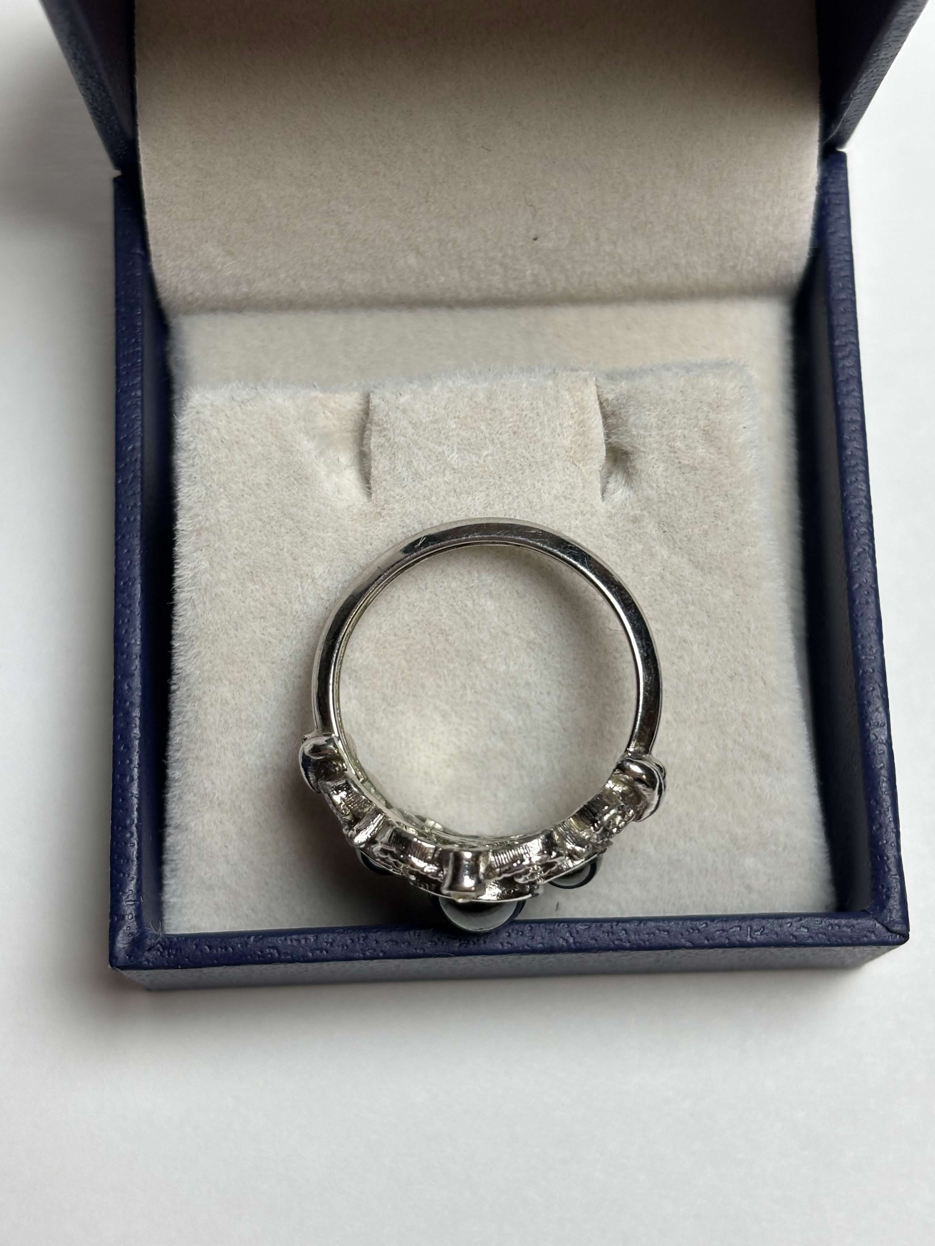 Side of silver tone ring with three oval black stones in the middle surrounded by filagree and has two clear stones at the highest points in a box on a neutral background