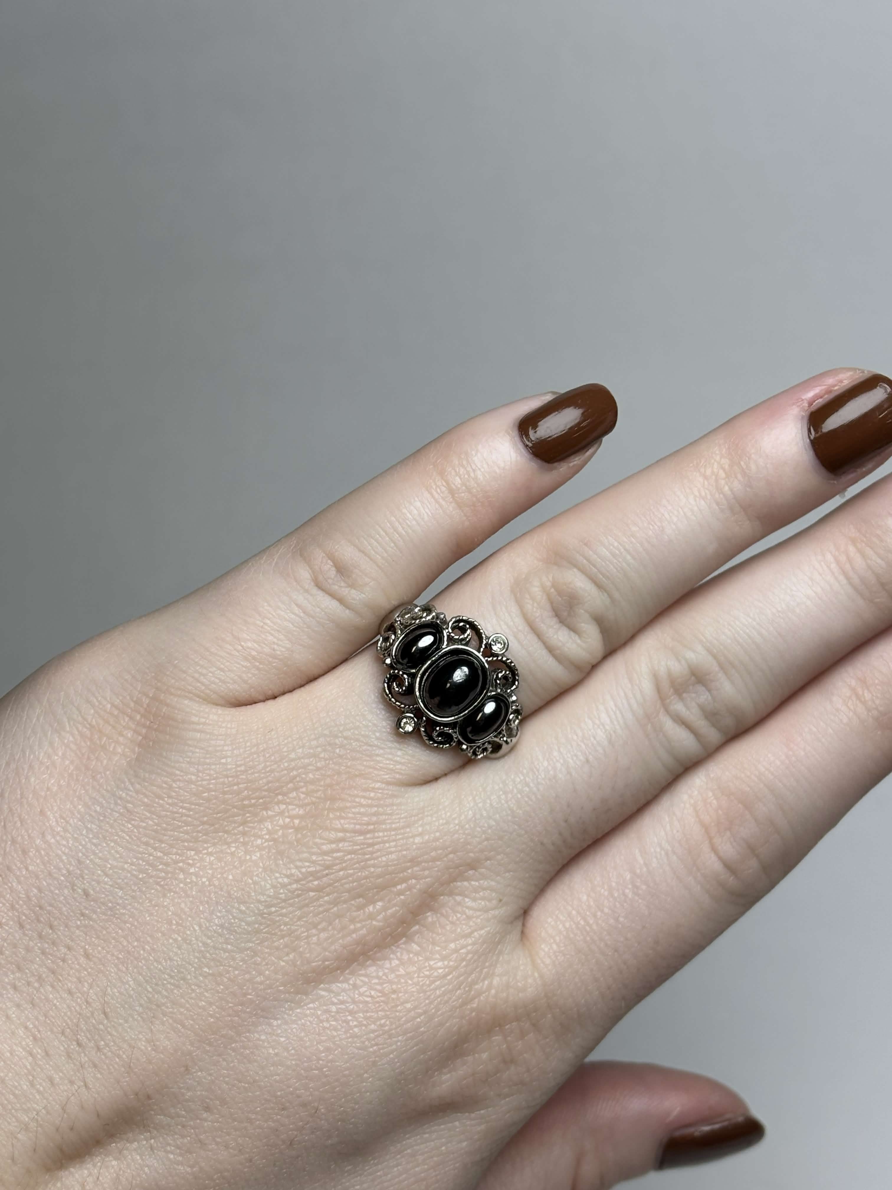 Front of silver tone ring with three oval black stones in the middle surrounded by filagree and has two clear stones at the highest points on a hand on a neutral background